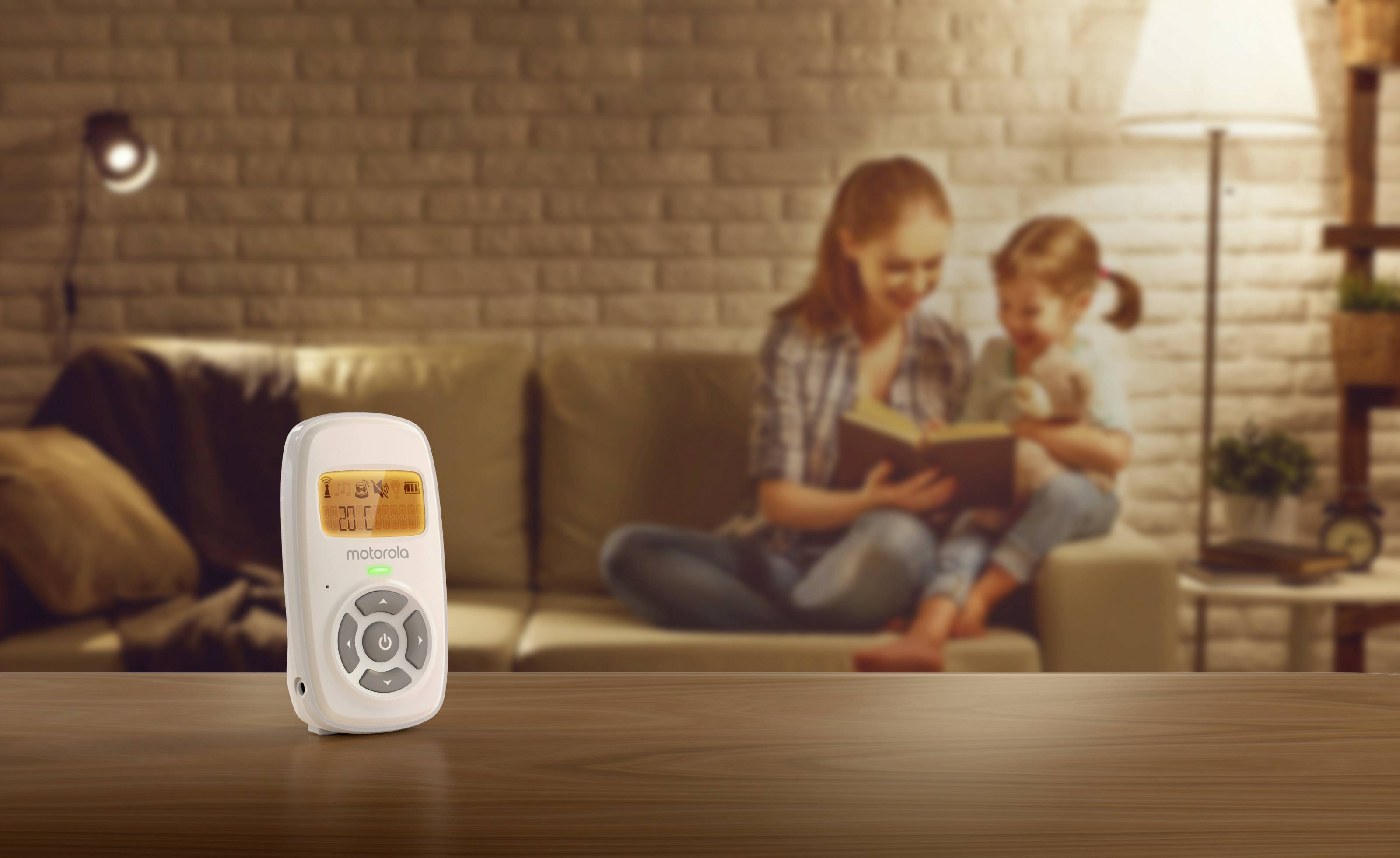 Ein Babyphone steht im Vordergrund auf einem Tisch, während eine Mutter im Hintergrund in einem warm beleuchteten Raum einem Kind vorliest.