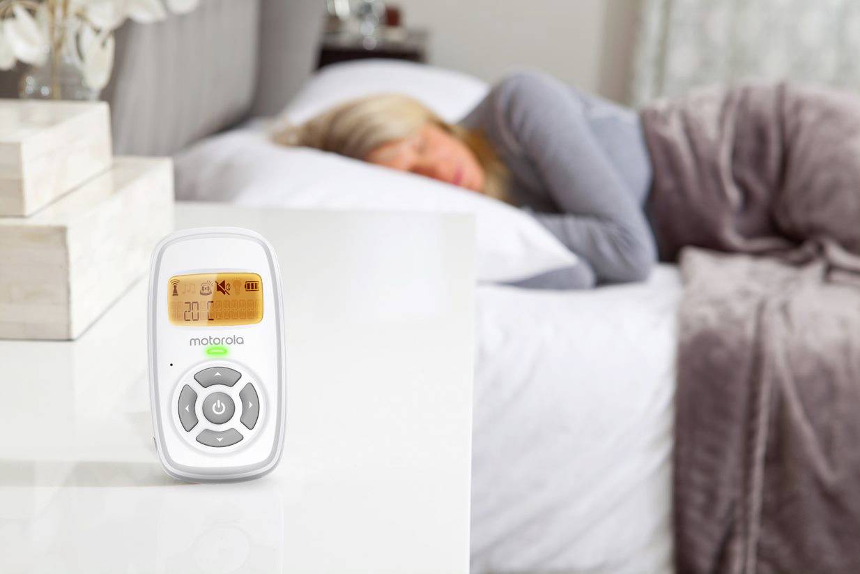 Eine Person schläft im Bett, während ein Babyphone auf einem nahe gelegenen Tisch steht und einen orangefarbenen Bildschirm mit Lautstärke- und anderen Funktionsreglern zeigt.