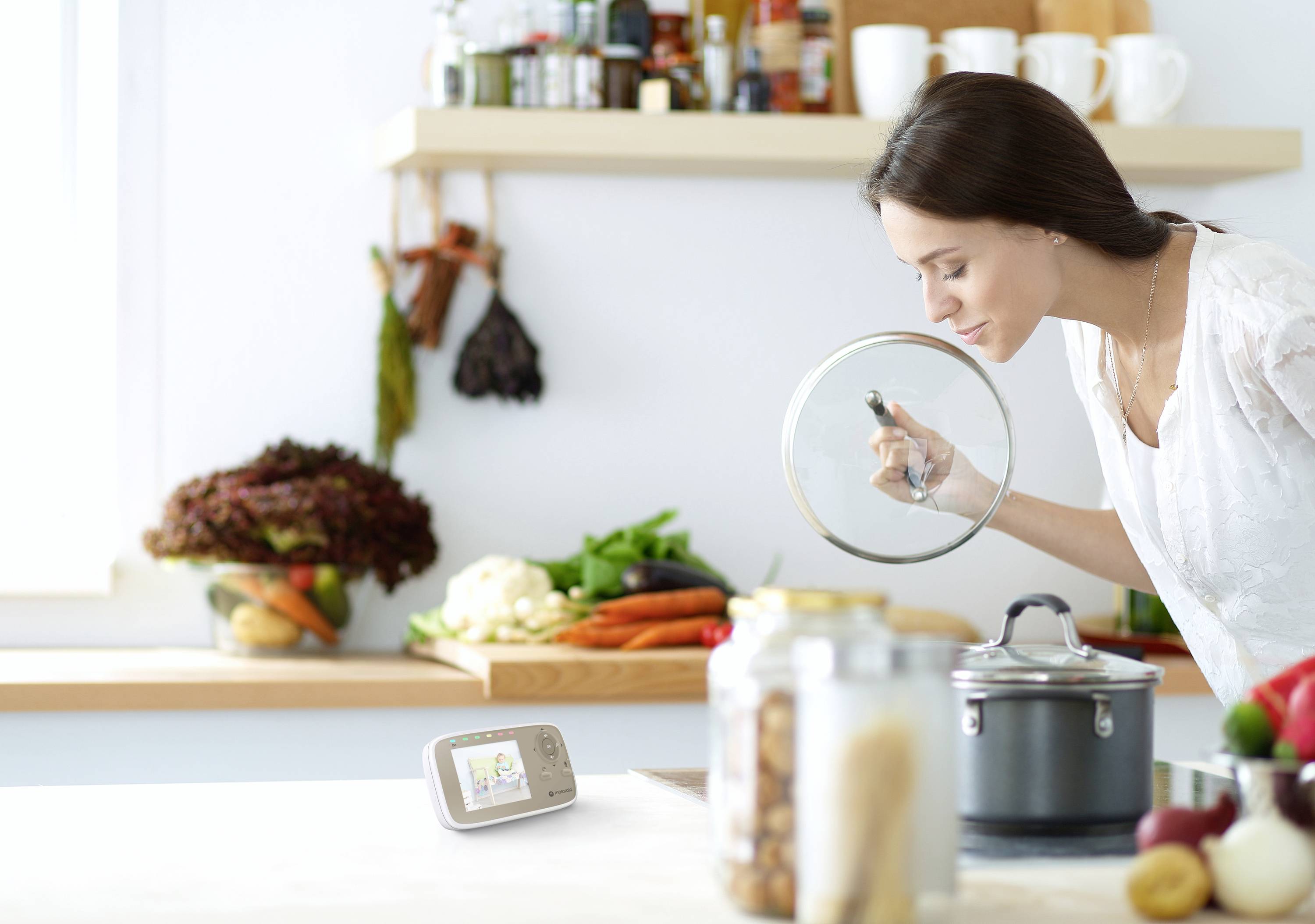 Eine Frau in einer Küche hebt einen Topfdeckel und prüft das Essen. Ein Babyphone auf der Arbeitsplatte zeigt ein Bild. Im Hintergrund befinden sich Gemüse und Gläser.