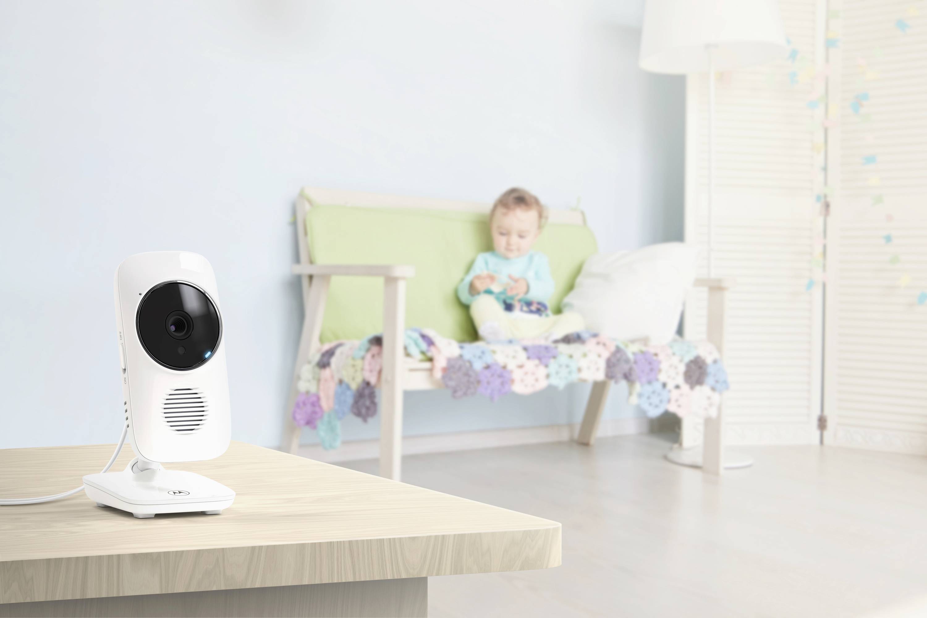 Ein Babyphone steht im Vordergrund auf einem Holztisch und erfasst ein Baby, das auf einem bunten Sofa in einem pastellfarbenen Raum sitzt.