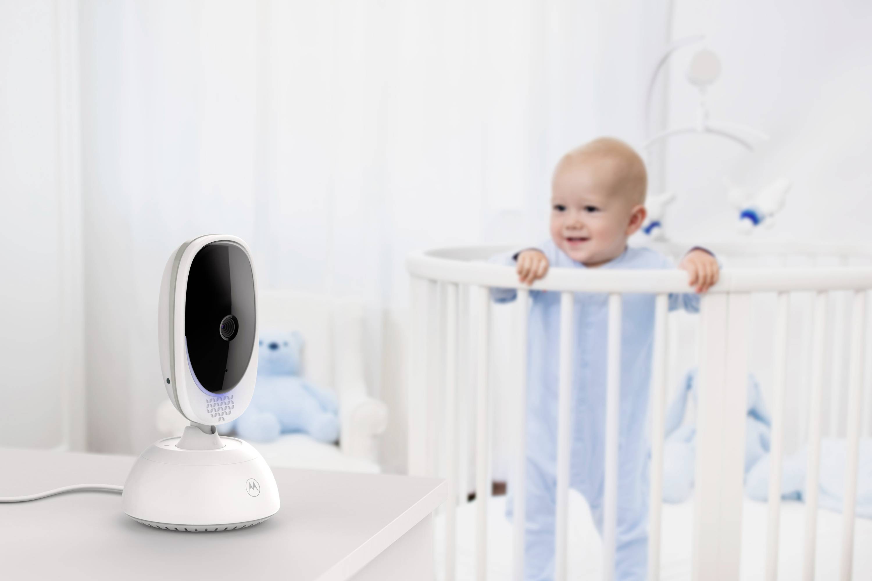 Ein Baby steht in einem Kinderbettchen in einem sanft beleuchteten Raum. Ein Babyphone liegt auf einem nahe gelegenen Tisch und ist auf das Kind gerichtet.