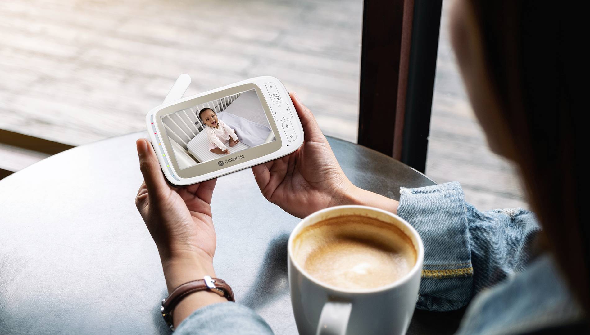 Eine Person hält einen Babyphone, auf dem ein schlafendes Baby zu sehen ist. Eine Tasse Kaffee steht auf dem Tisch und deutet auf eine entspannte Häuslichkeit hin.