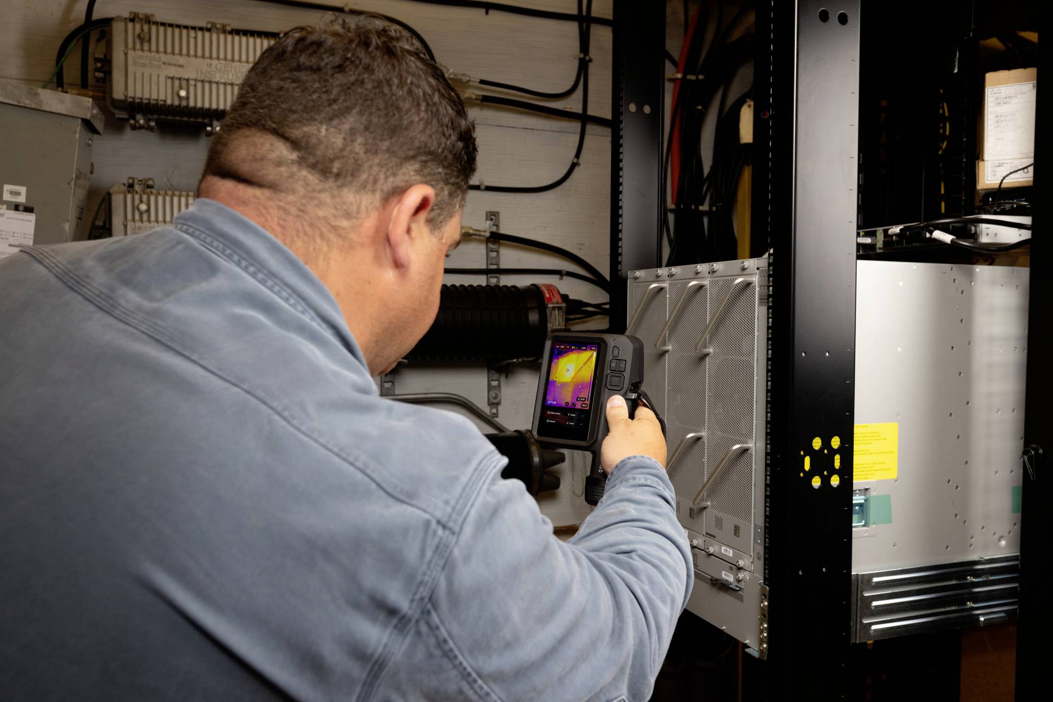 Eine Person in einer Arbeitsjacke verwendet eine Wärmebildkamera zur Inspektion von elektrischen Geräten in einer industriellen Umgebung.