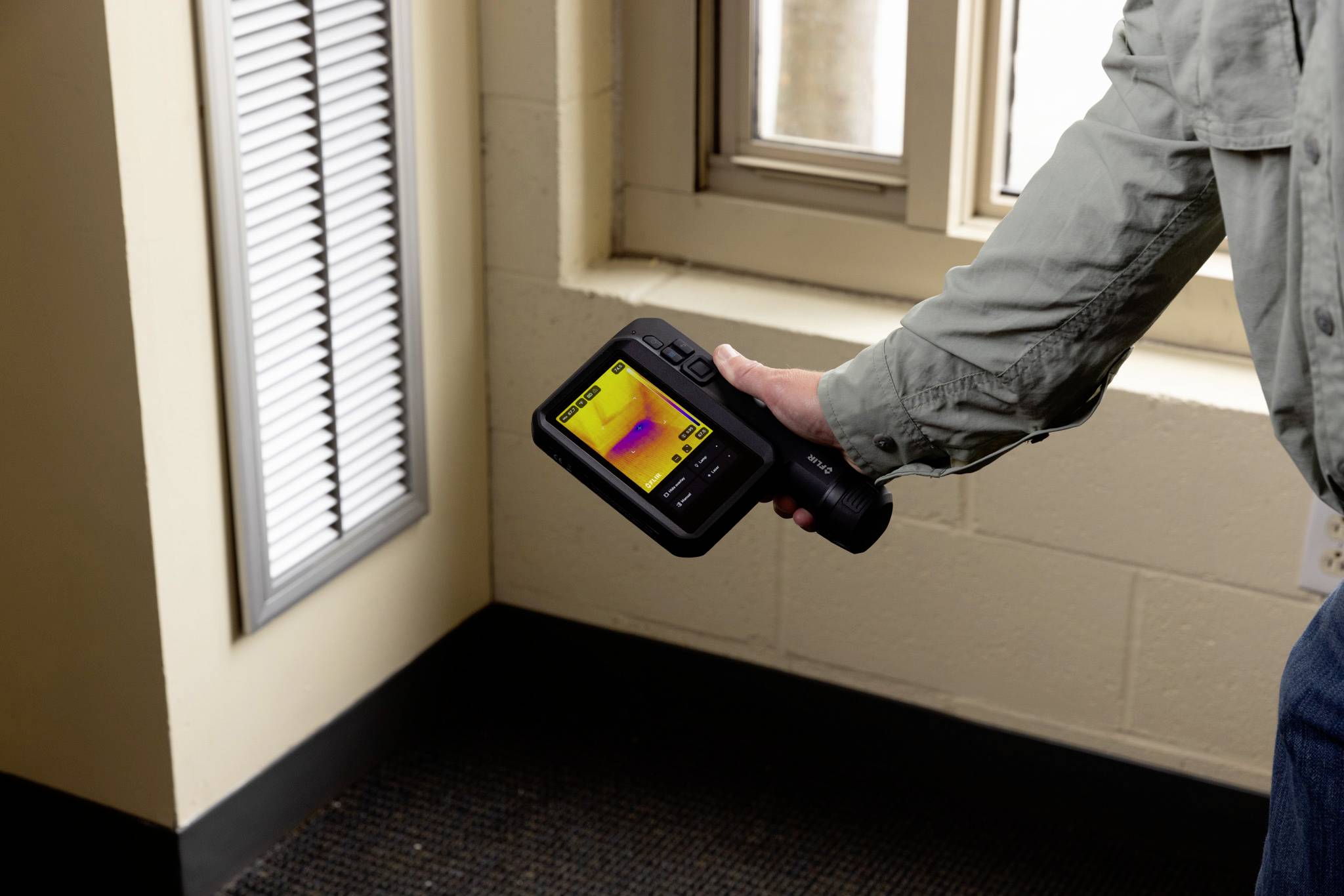 Person hält eine Wärmebildkamera neben einer Wandlüftung und zeigt eine Wärmekarte auf dem Bildschirm, die Temperaturunterschiede anzeigt.