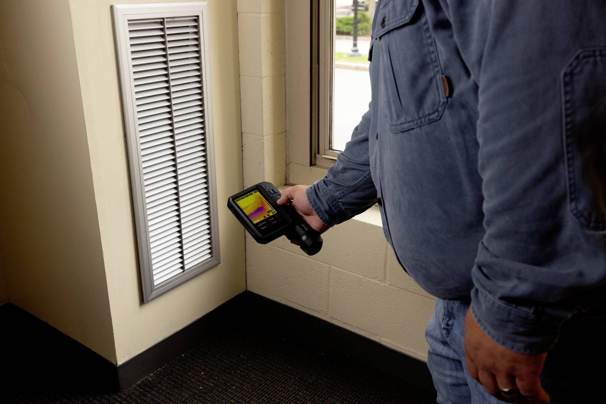 Eine Person, die eine tragbare Wärmekamera verwendet, um die Temperatur an einem Wandlüftungsschacht im Innenraum zu überprüfen und dabei Wärmeverluste oder Isolationsprobleme untersucht.