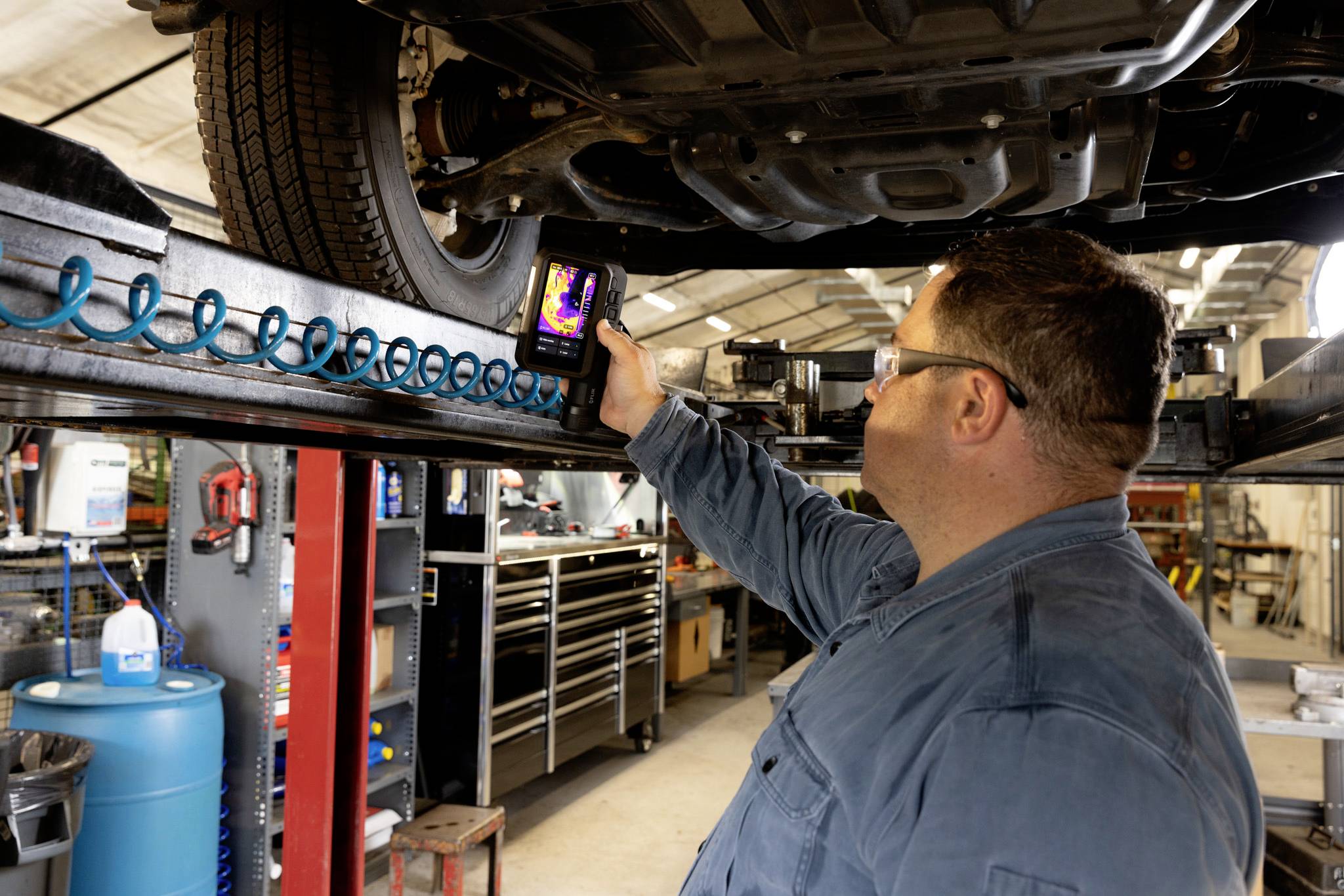 Ein Mechaniker in einer Werkstatt verwendet eine Wärmebildkamera, um den Untergrund eines Fahrzeugs zu überprüfen und konzentriert sich dabei auf einen Reifen. Werkzeuge und Ausrüstung sind zu sehen.