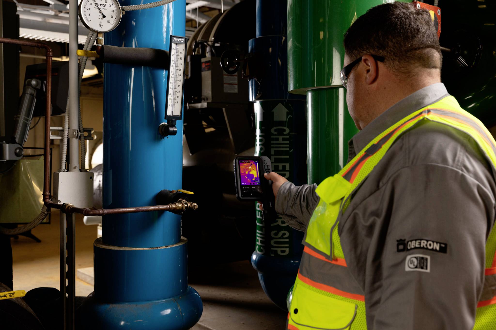 Eine Person in Schutzausrüstung verwendet eine Infrarotkamera, um die Temperaturen an industriellen Rohrleitungen in einem Maschinenraum zu überprüfen.
