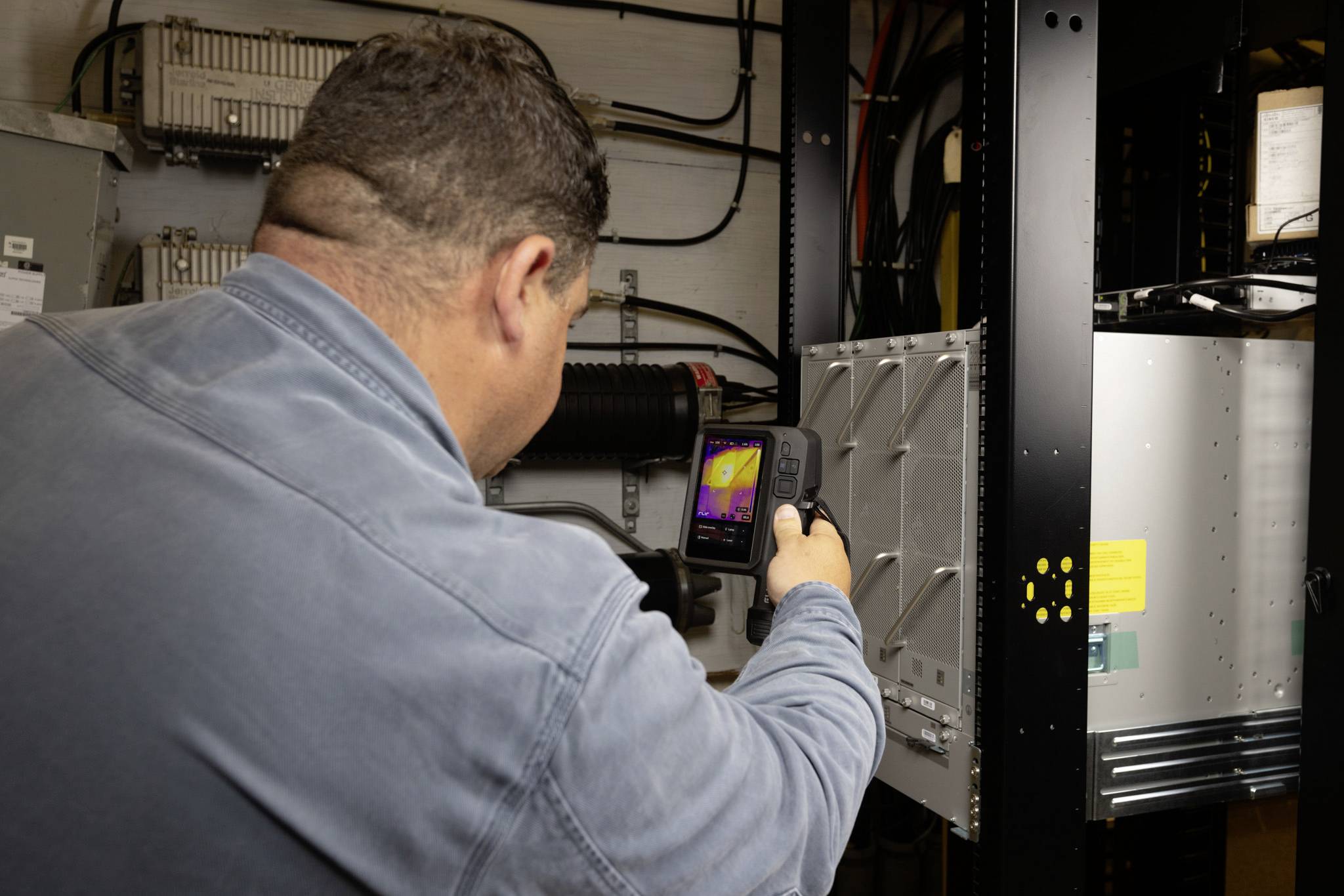 Eine Person in einer blauen Jacke verwendet eine Wärmebildkamera zur Inspektion von elektronischer Ausrüstung in einem technischen Raum.
