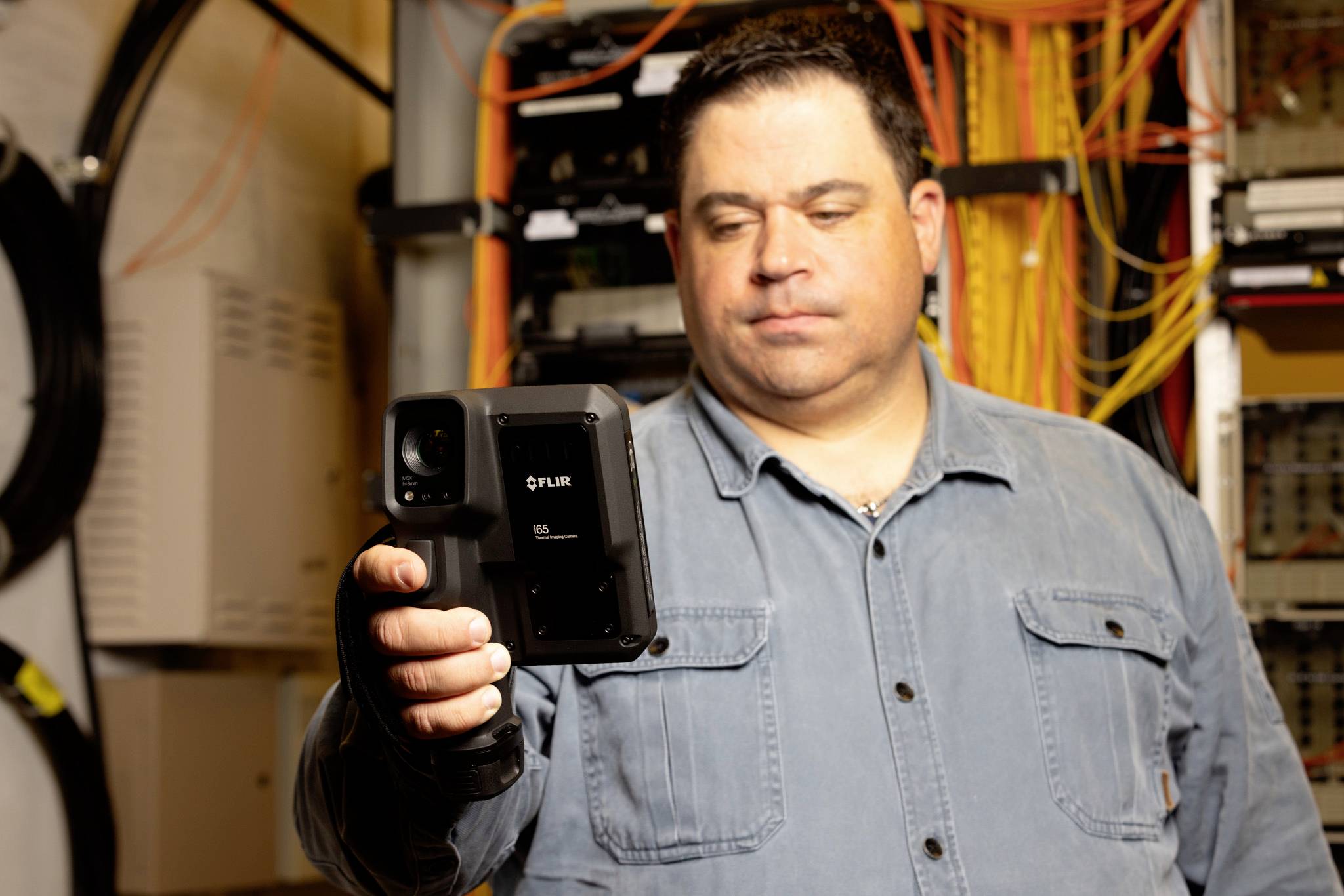 Eine Person hält eine FLIR-Wärmebildkamera in einem Serverraum, wobei Kabel und Ausrüstung im Hintergrund auf eine technische Umgebung hinweisen.