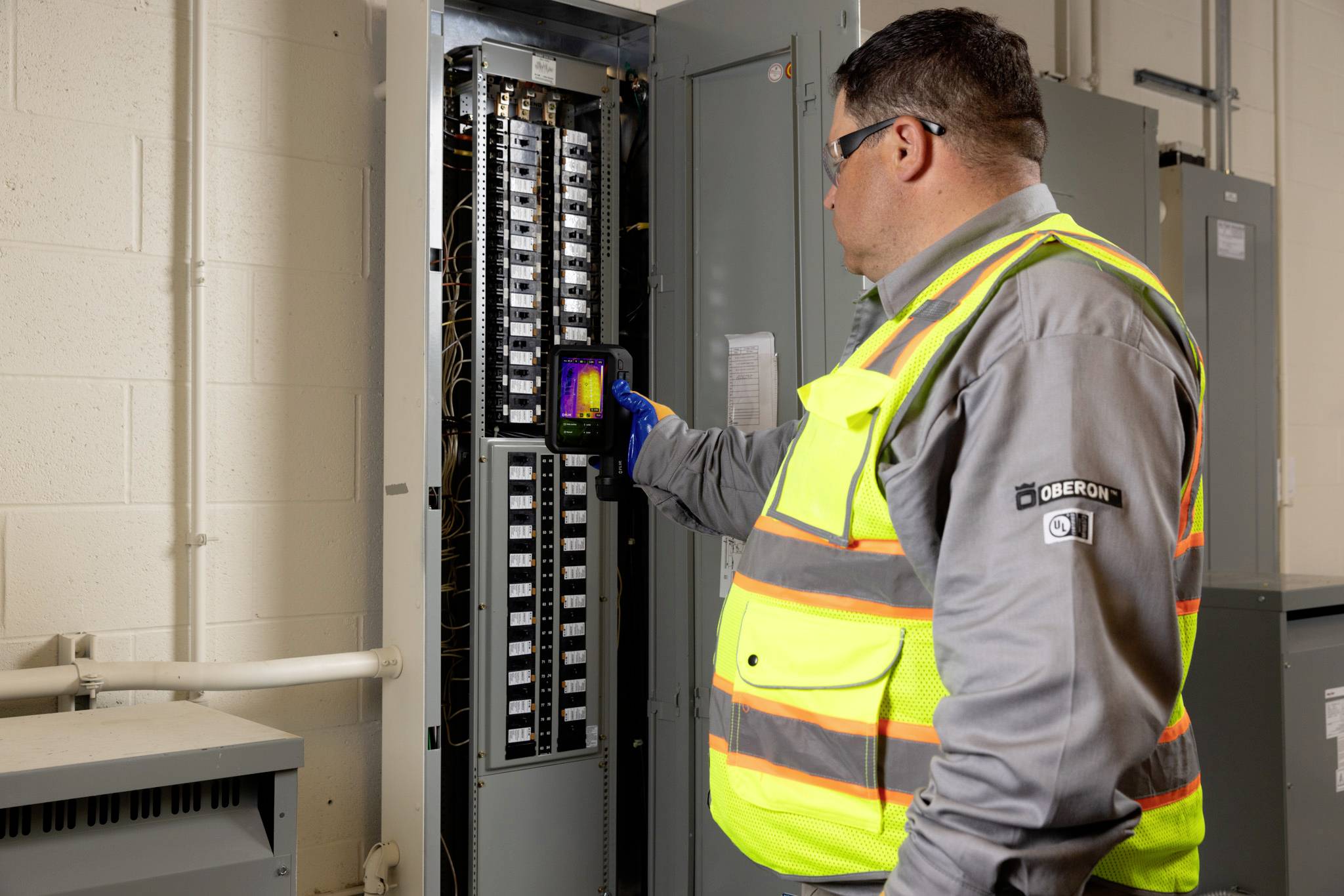 Eine Person mit Sicherheitsweste und Schutzbrille verwendet eine tragbare Infrarot-Wärmekamera zur Inspektion einer offenen Elektrotafel in einem Versorgungsraum.
