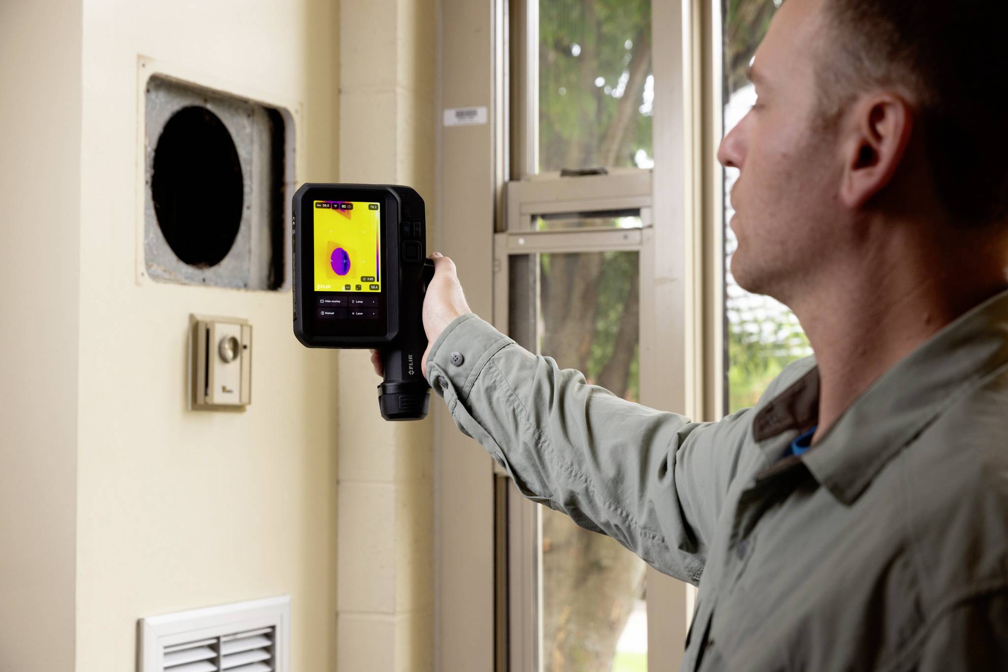 Eine Person verwendet eine Wärmebildkamera in der Nähe eines Lüftungsschachts und zeigt Temperaturunterschiede in verschiedenen Farben auf dem Bildschirm.
