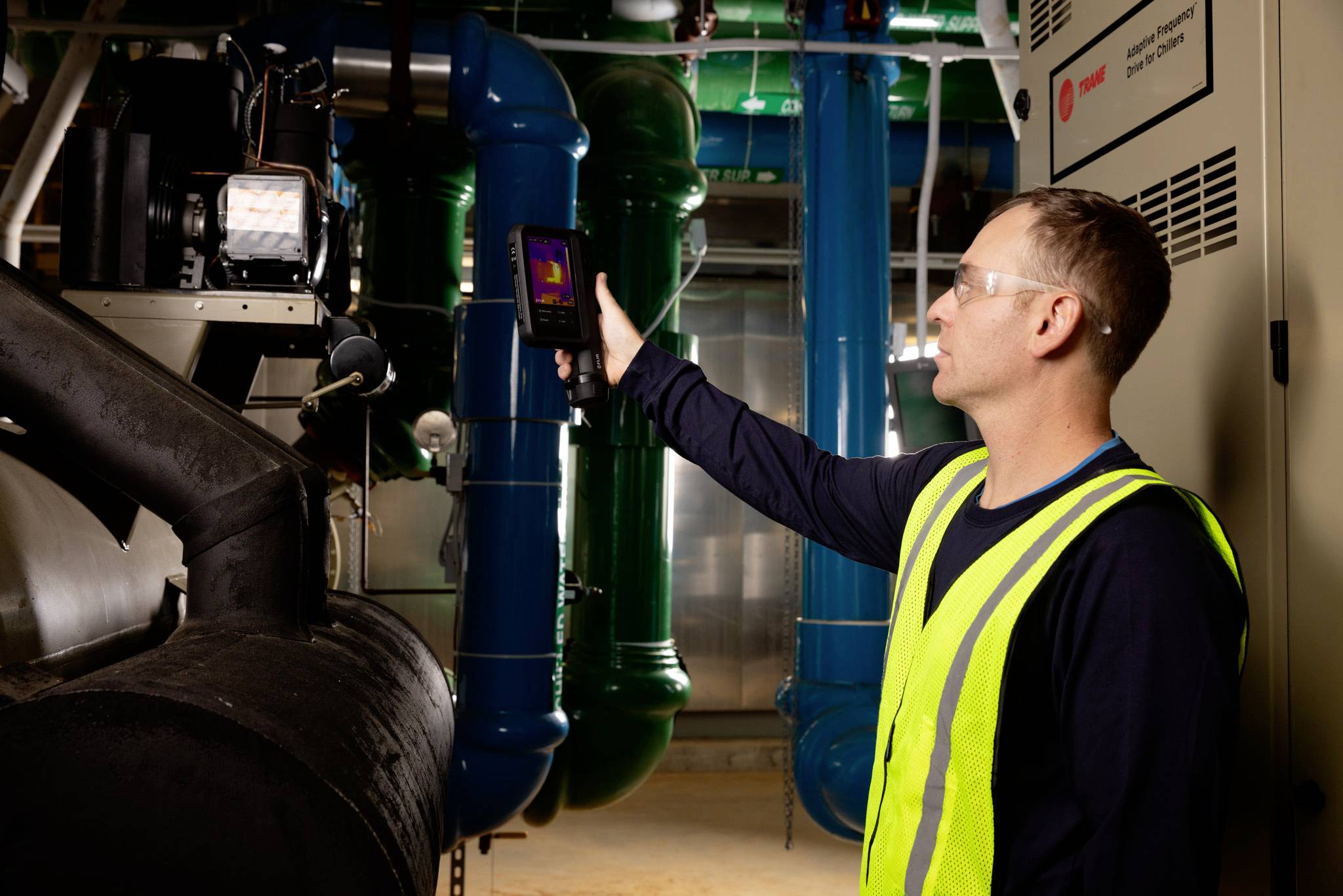 Ein Techniker in einer Sicherheitsweste verwendet eine Wärmebildkamera zur Inspektion von industriellen Rohrleitungen in einem Maschinenraum und gewährleistet dabei die Sicherheit der Ausrüstung.