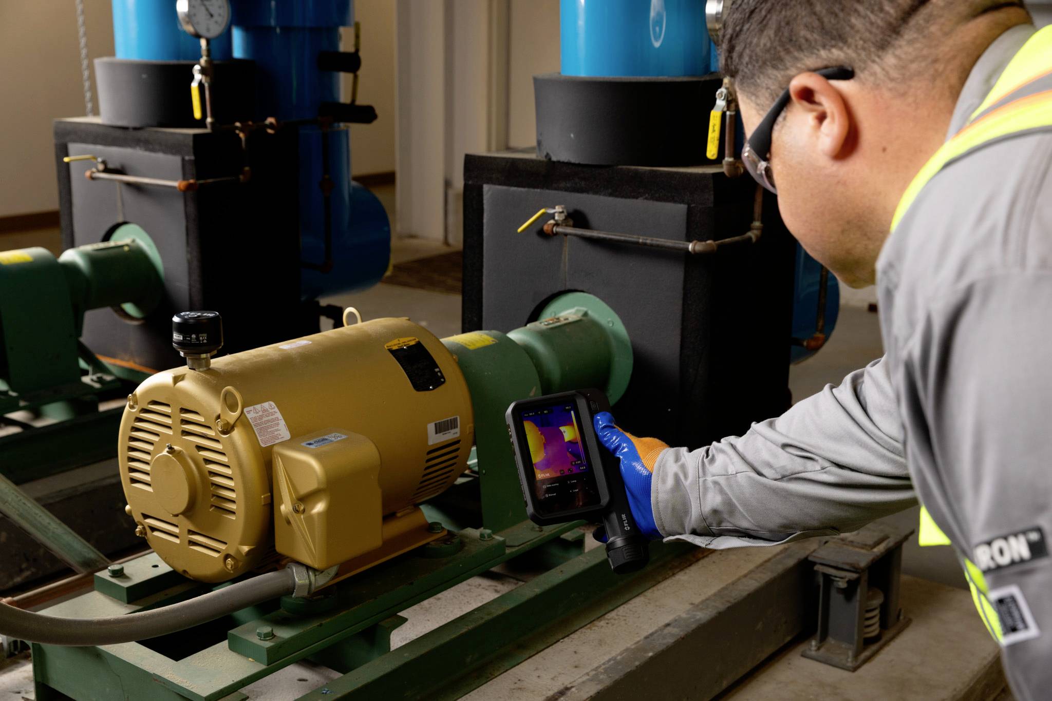 Ein Techniker in Sicherheitsausrüstung verwendet eine Wärmebildkamera zur Inspektion von industrieller Maschinerie und konzentriert sich dabei auf einen großen Motor und Rohrleitungen.