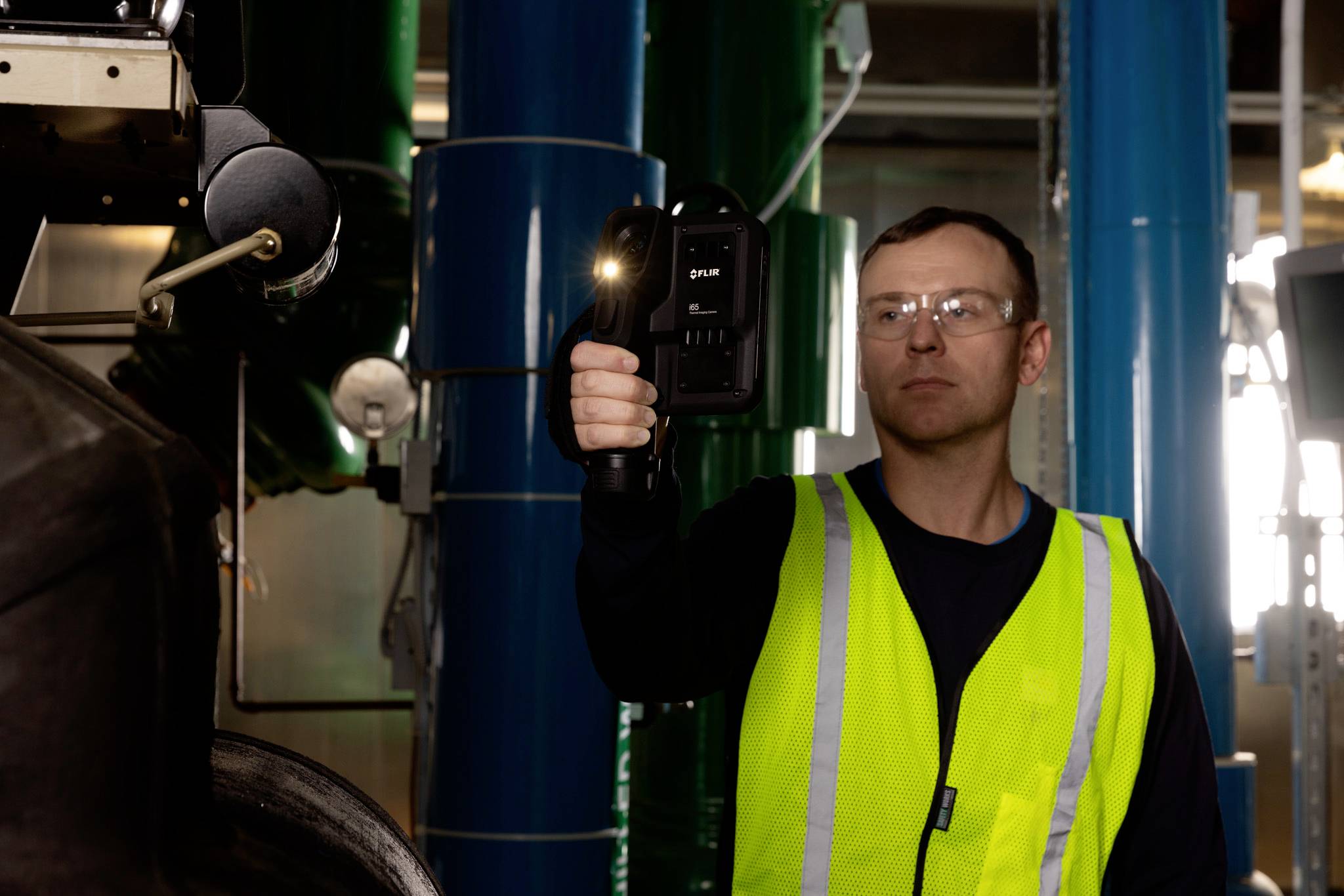 Ein Mann in einer Warnweste bedient ein Handgerät zur Inspektion von Maschinen in einer industriellen Umgebung, wobei blaue Rohre im Hintergrund zu sehen sind.