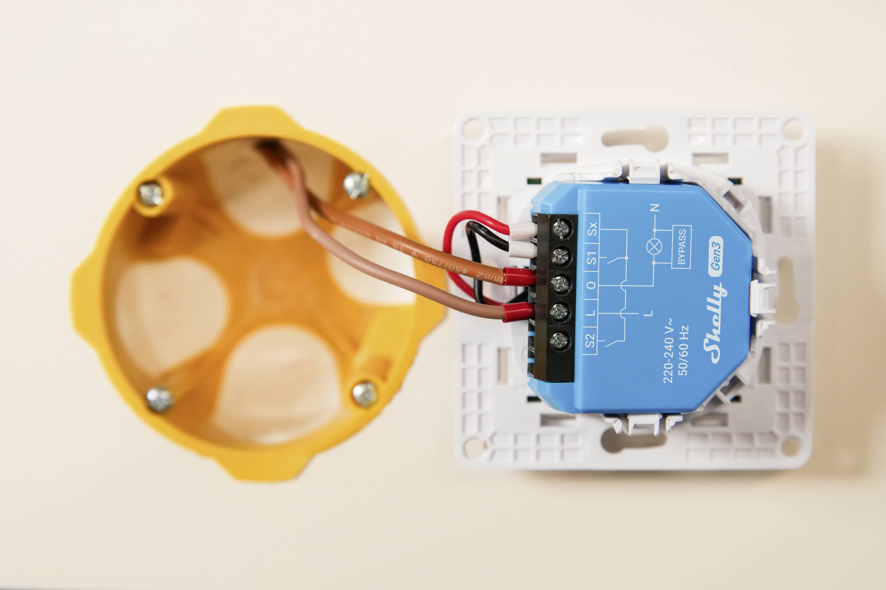 Ein blaues Shelly-Schaltmodul, das mit Kabeln neben einer orangefarbenen Elektro-Verteilerdose auf beigem Hintergrund verbunden ist.