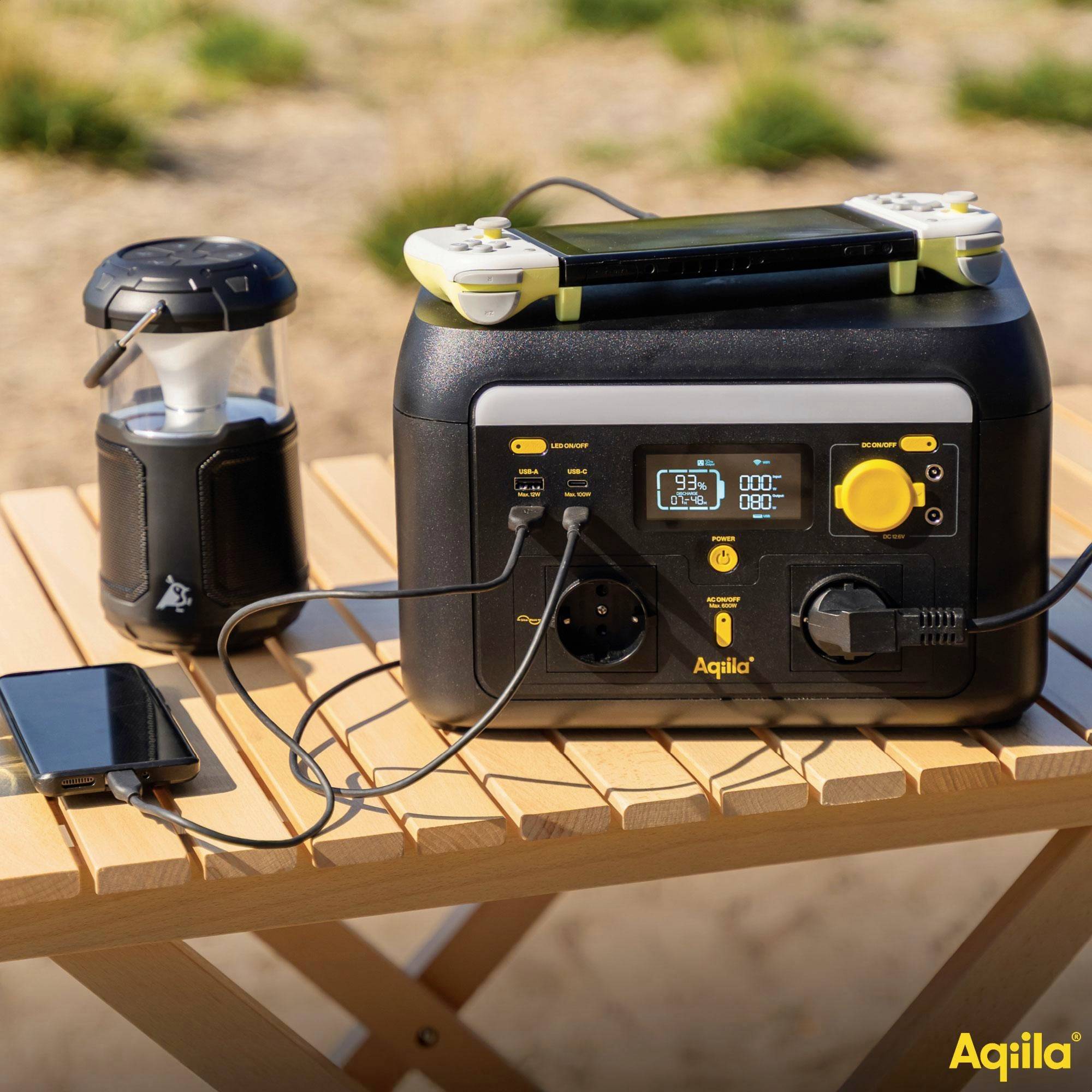 Eine mobile Powerstation lädt ein Handy und eine Spielkonsole im Freien. Eine Laterne steht in der Nähe auf einem Holztisch. Logo: 'Aqiiila'.
