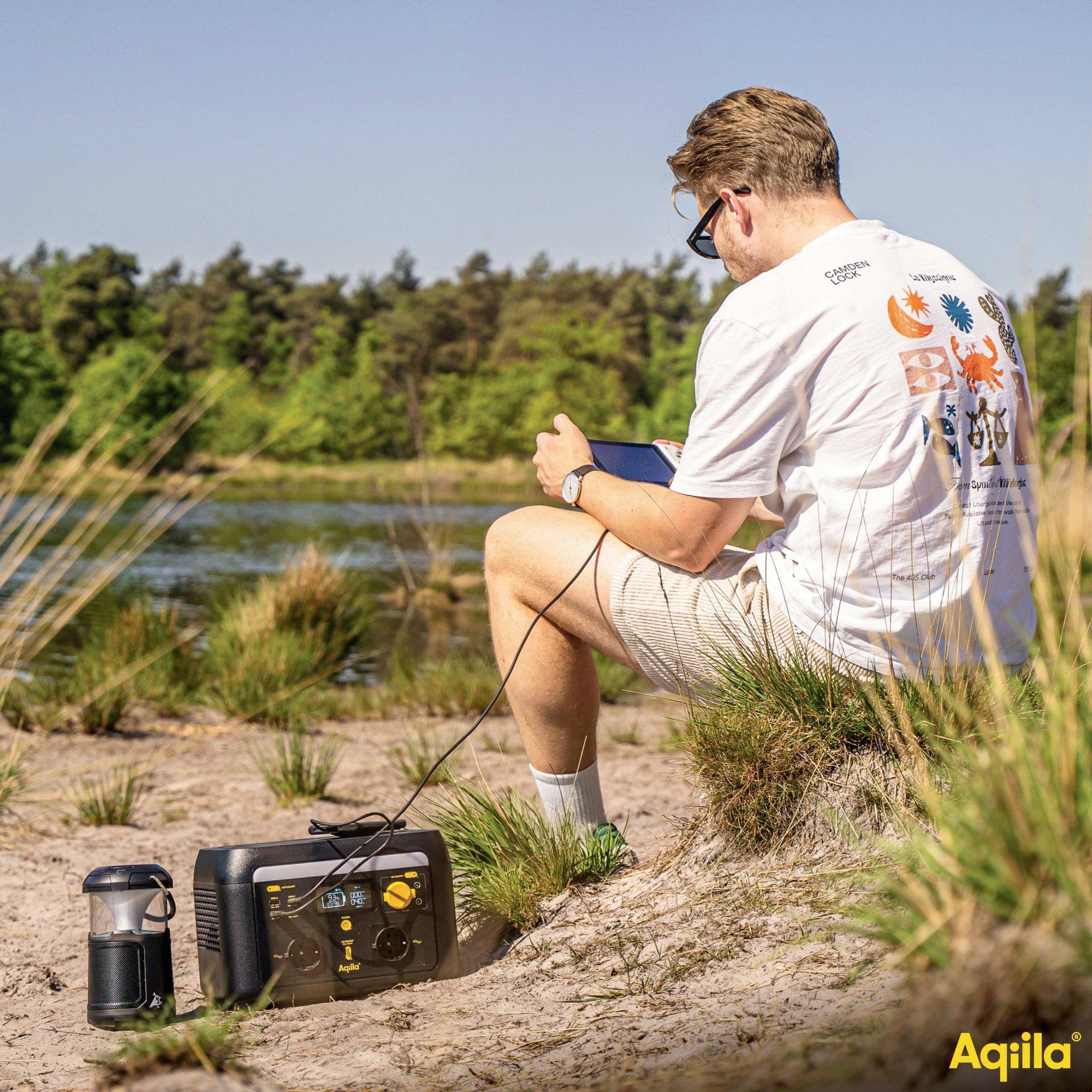 Eine Person sitzt im Freien und bedient elektronische Geräte, die an eine mobile Stromversorgungsstation angeschlossen sind, während im Hintergrund eine natürliche Landschaft zu sehen ist.