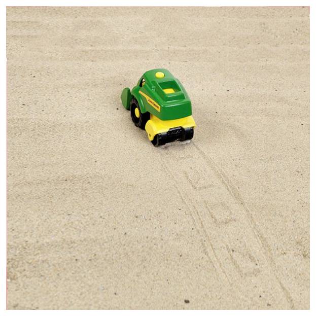 Ein kleiner Spielzeugtraktor, grün und gelb, bewegt sich über Sand und hinterlässt Spuren.