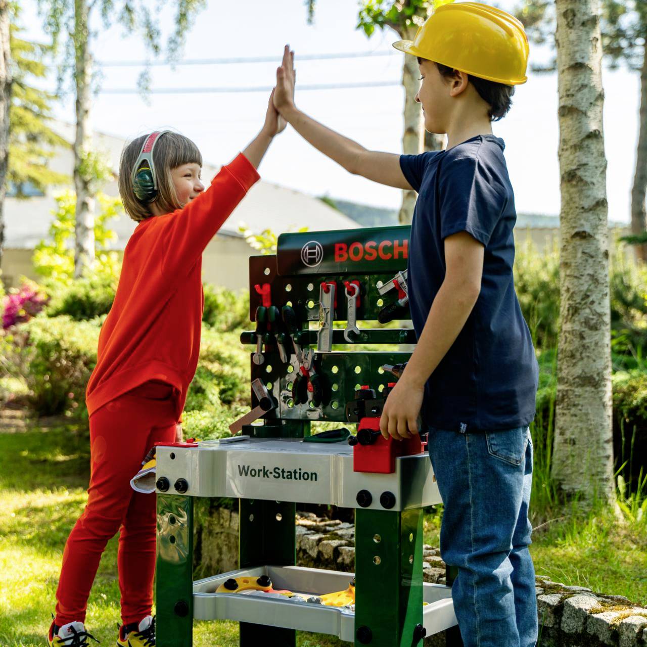 Ein Kind mit Schutzhelm und ein anderes mit Gehörschutz geben sich draußen an einer Spielwerkelbank ein High-Five, was Teamarbeit und Spielfreude symbolisiert.