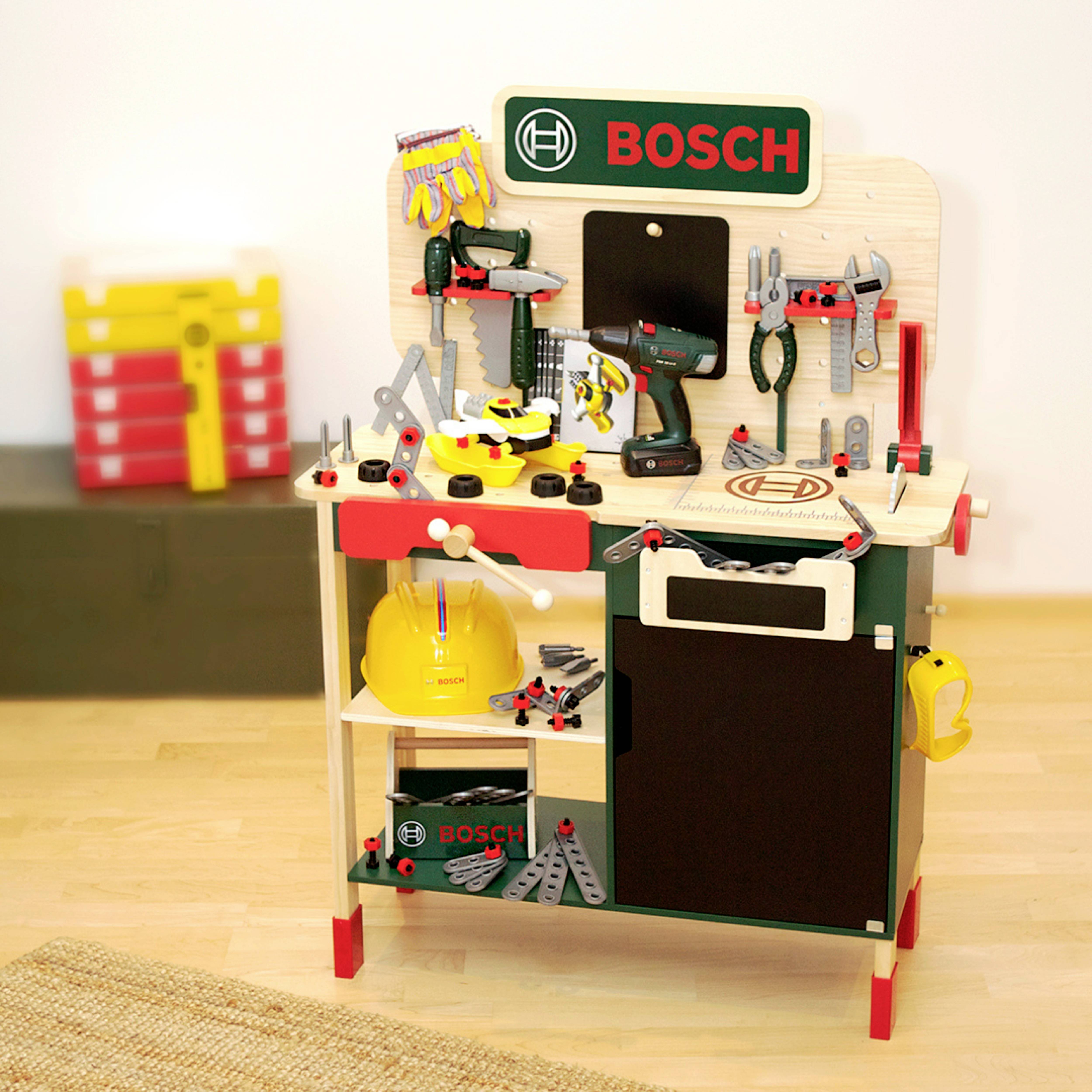Eine Kinder-Werkbank mit verschiedenen Spielwerkzeugen und -zubehör, mit der Marke 'Bosch' versehen, und einem gelben Spielzeug-Schutzhelm auf einem Regal.