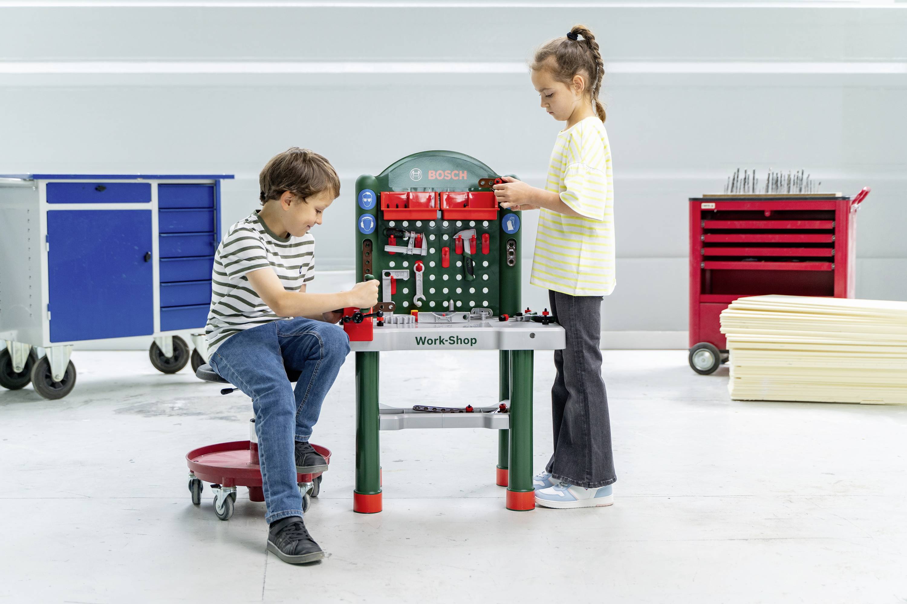 Ein Junge und ein Mädchen spielen mit einer Bosch-Spielzeug-Werkbank und ordnen Werkzeuge an. Ein roter und blauer Werkzeugwagen befindet sich im Hintergrund.