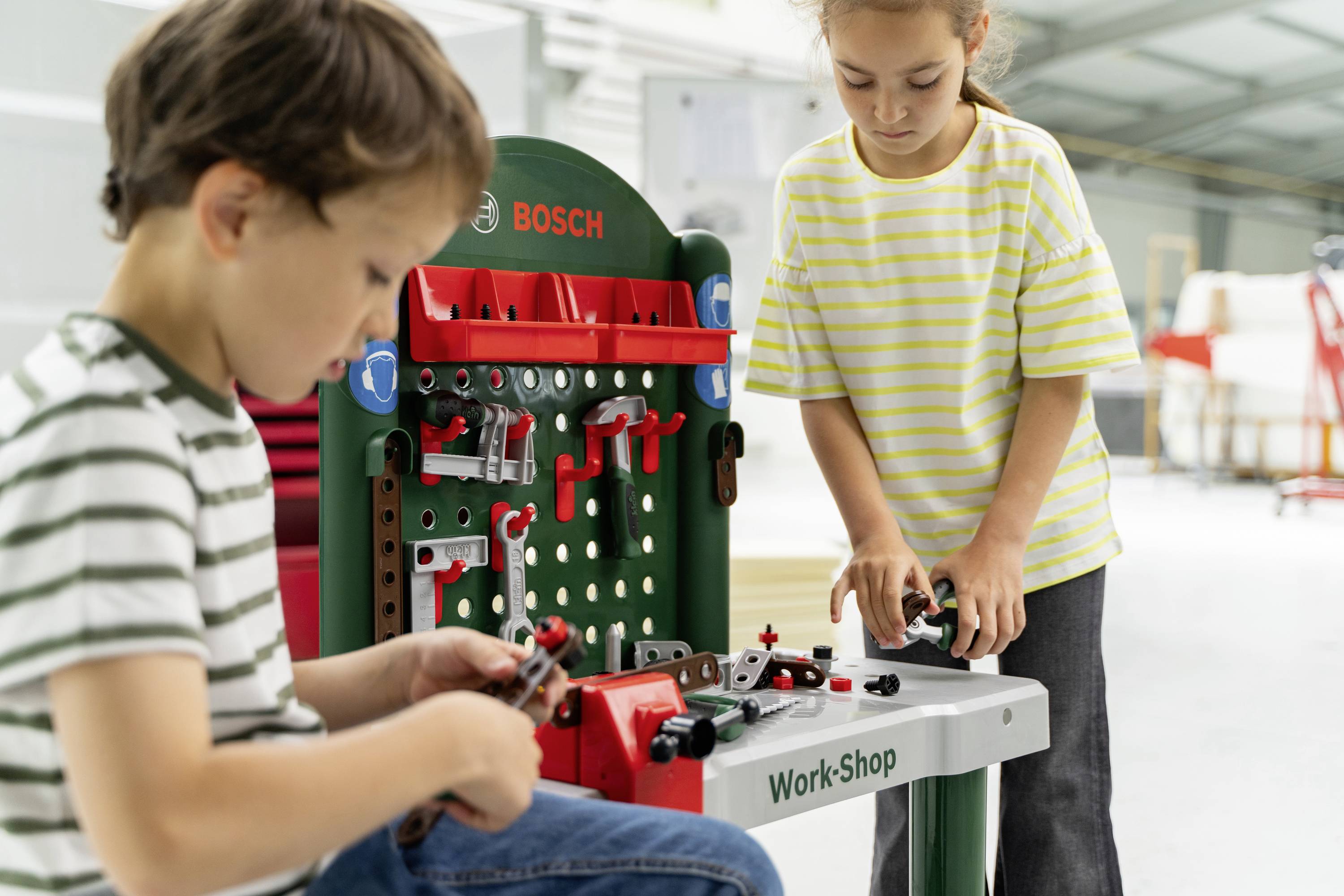 Zwei Kinder spielen drinnen mit einem Bosch Spielzeug-Werkstattset und nutzen verschiedene Spielzeugwerkzeuge und -teile.