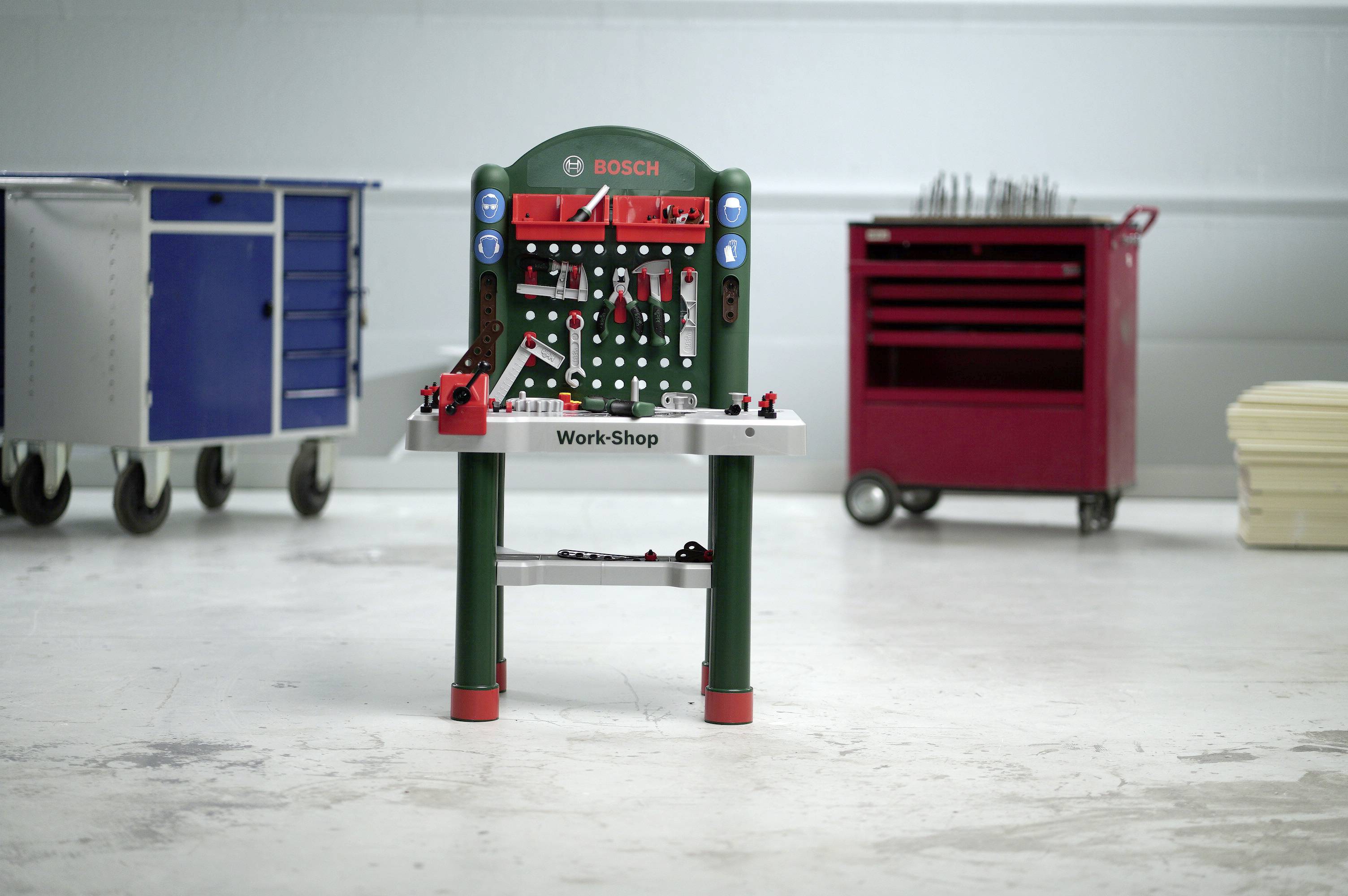 Eine Spielzeug-Werkbank mit dem Etikett 'BOSCH' und 'Work-Shop' steht in einer Garage. Im Hintergrund sind zwei Werkzeugschränke zu sehen.