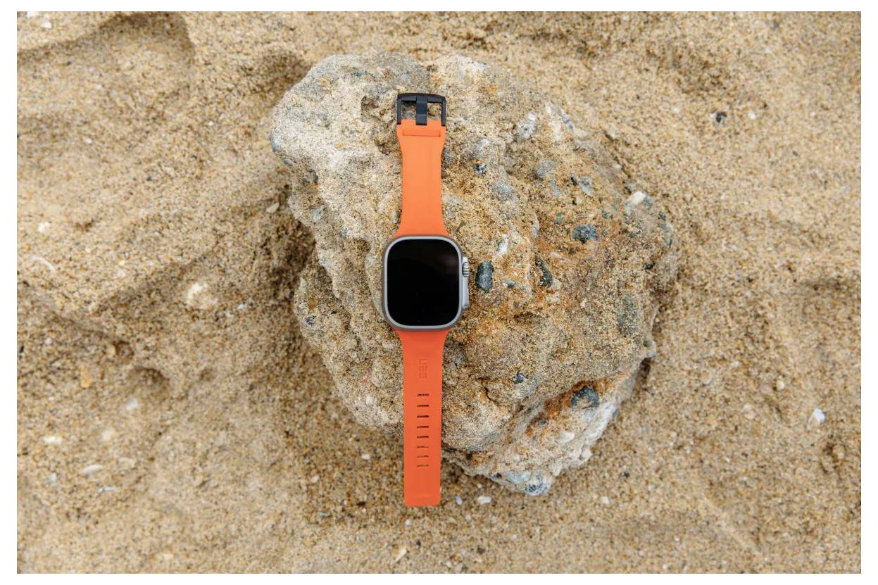 Eine Smartwatch mit einem orangefarbenen Armband liegt auf einem Stein in einem sandigen Gebiet.