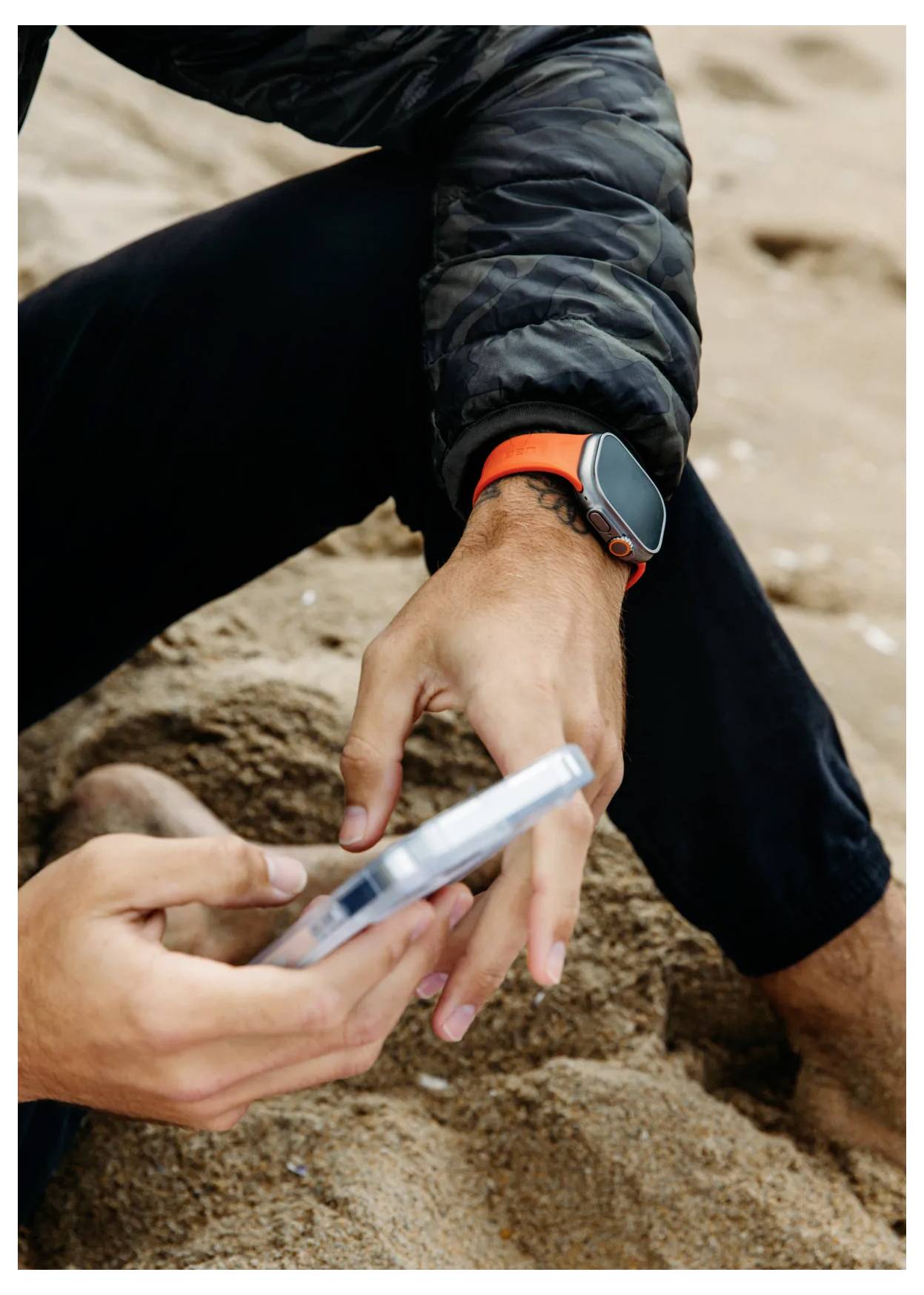 Eine Person, die auf dem Sand sitzt und ein Smartphone benutzt, trägt eine Jacke und eine Smartwatch mit einem orangefarbenen Armband.