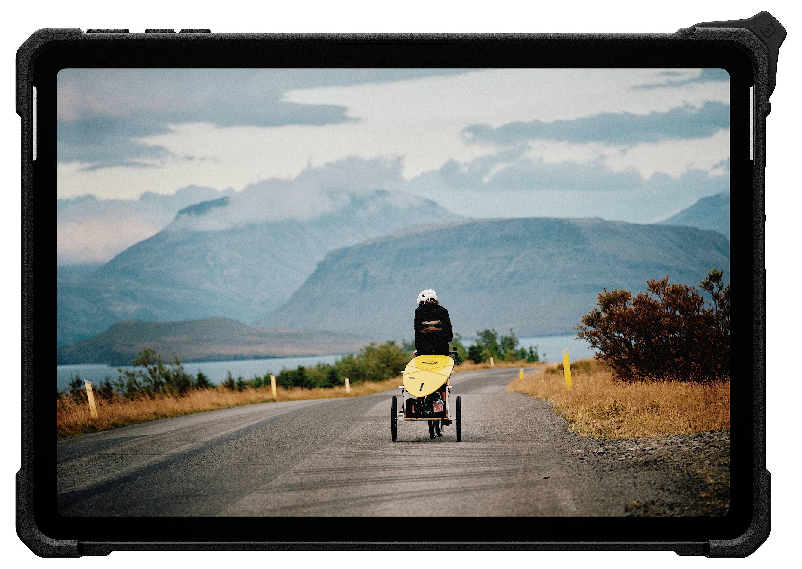 Eine Person fährt auf einem gelben Liegerad eine ländliche Straße entlang in Richtung ferner Berge unter einem bewölkten Himmel.