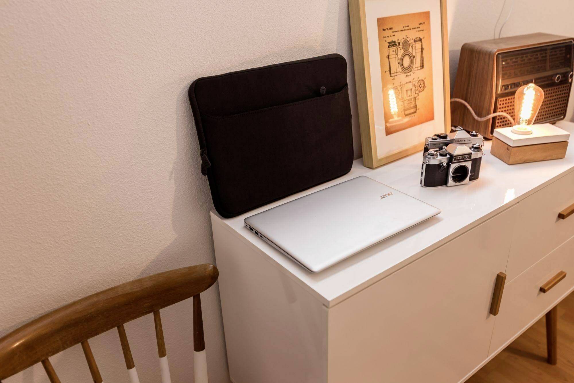 Ein kleiner weißer Schrank mit einem geschlossenen silbernen Laptop, einer Vintage-Kamera und einem gerahmten Sketch obenauf. Retroradio mit warm leuchtenden Glühbirnen in der Nähe.