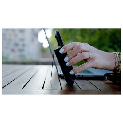 Eine Hand mit hellblauen Nägeln und Armbändern berührt ein Smartphone auf einem Holztisch im Freien, während ein Laptop im Hintergrund zu sehen ist.