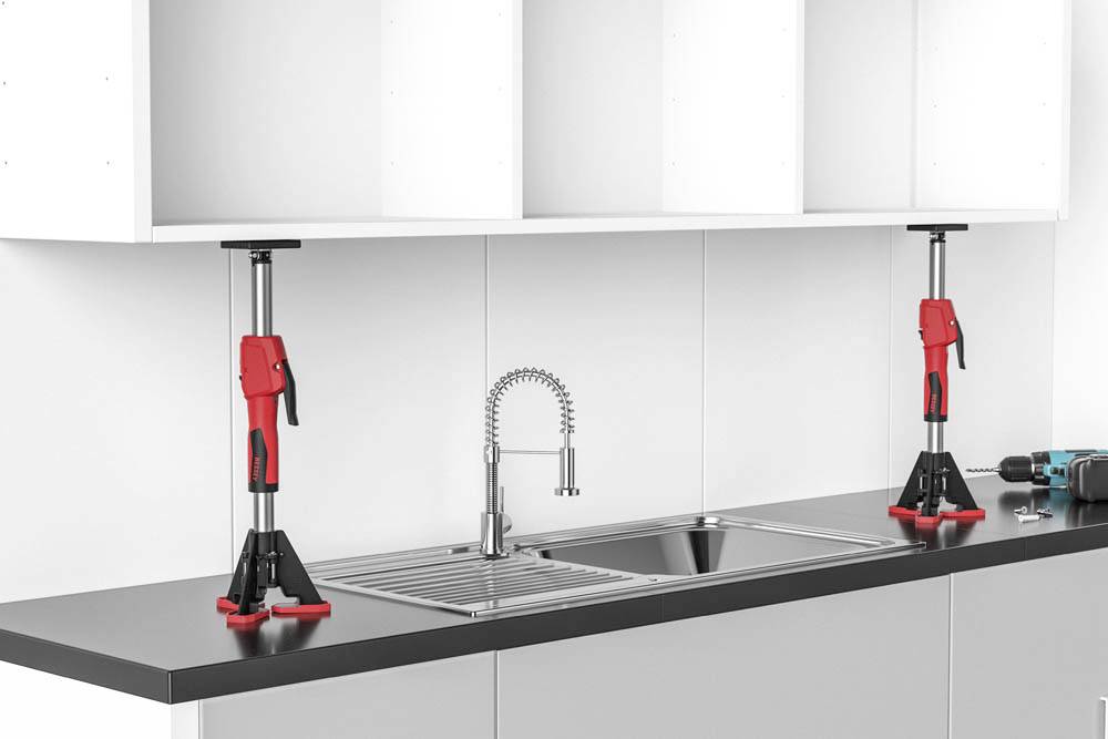 Zwei rote Schrankhebevorrichtungen stützen einen weißen Hängeschrank über einem Waschbecken mit Wasserhahn, wobei auf der Arbeitsplatte in der Nähe eine Bohrmaschine liegt.