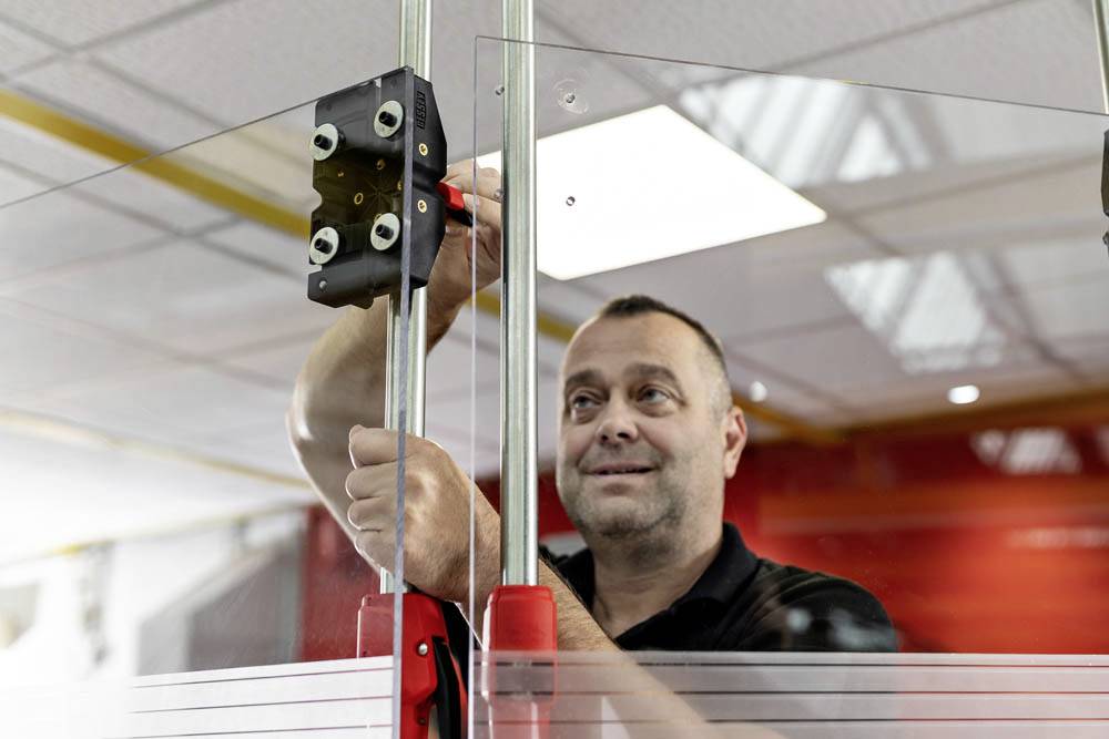 Eine Person justiert eine transparente Schutzbarriere mit Metallstangen und Halterungen in einem Büroumfeld und konzentriert sich dabei auf Sicherheit und Installation.