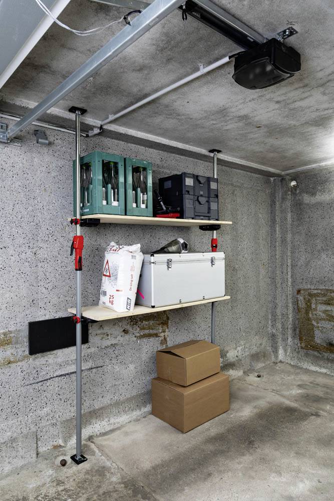 Regale in einer Garage halten Werkzeugkästen und Materialien. Zwei Kisten sind unten auf dem Betonboden gestapelt. Die Wände sind aus blankem Beton.