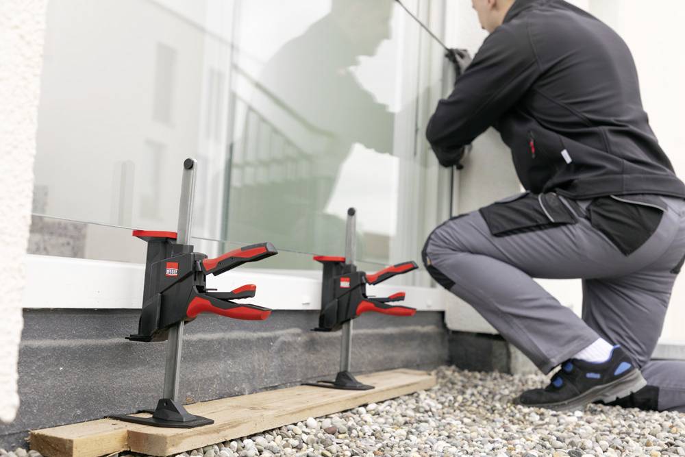 Eine Person verwendet Klemmen, um ein großes Glasfenster an der Außenfassade eines Gebäudes zu installieren. Zwei Klemmen sind auf einem Holzbrett zur Stabilisierung angebracht.