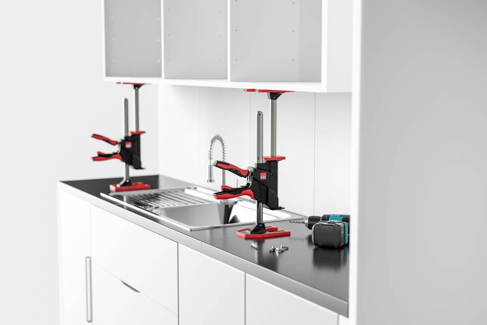 Küchenarbeitsplatte mit einer Szene, in der zwei rot-schwarze Schraubzwingen einen Schrank sichern. In der Nähe liegen eine Bohrmaschine, ein Schraubendreher und Schrauben auf der Arbeitsplatte.
