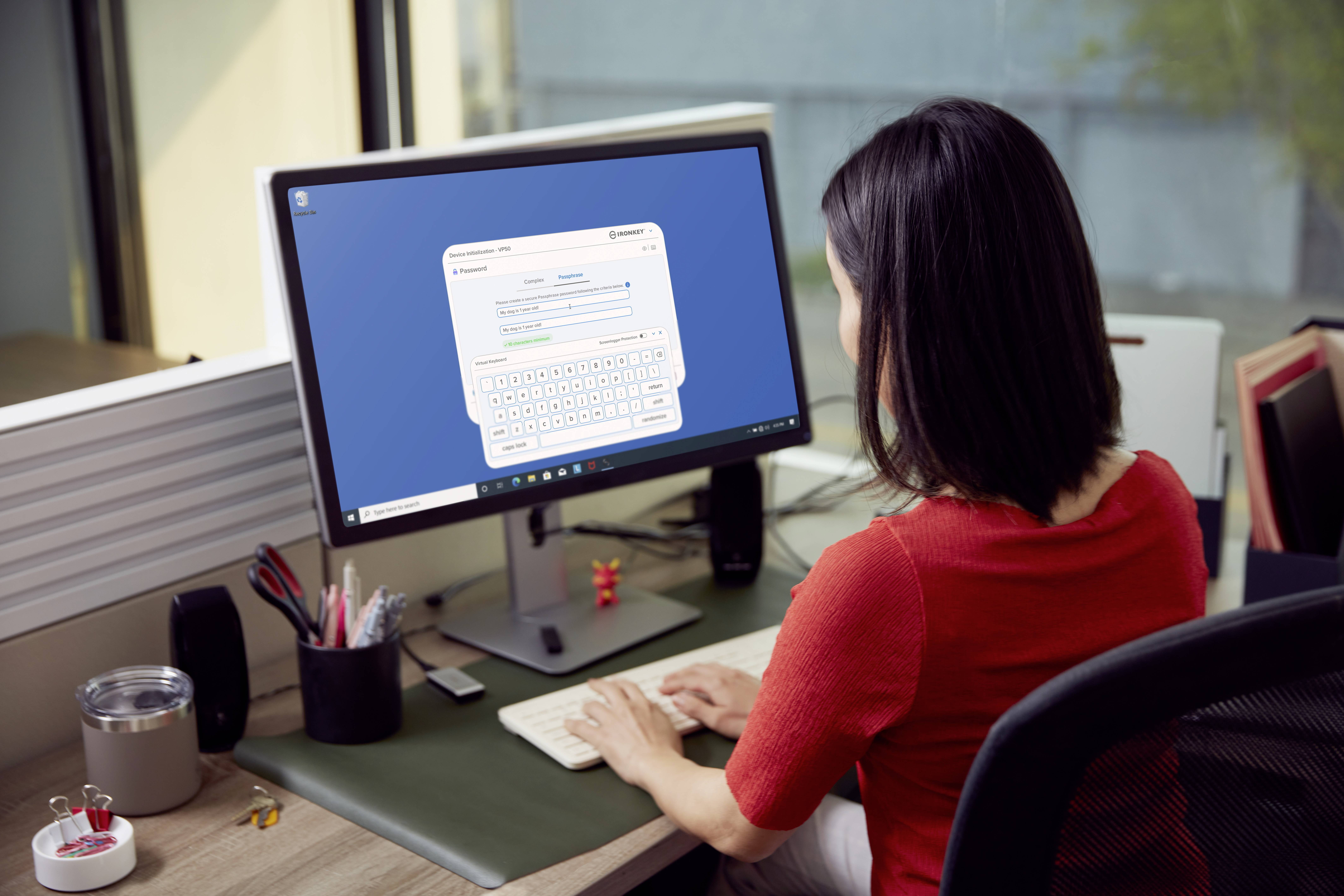 Eine Frau in einem roten Hemd sitzt an einem Schreibtisch und arbeitet an einem Computer mit einem großen Monitor, auf dem eine Tastatur und Texteingabe zu sehen sind.