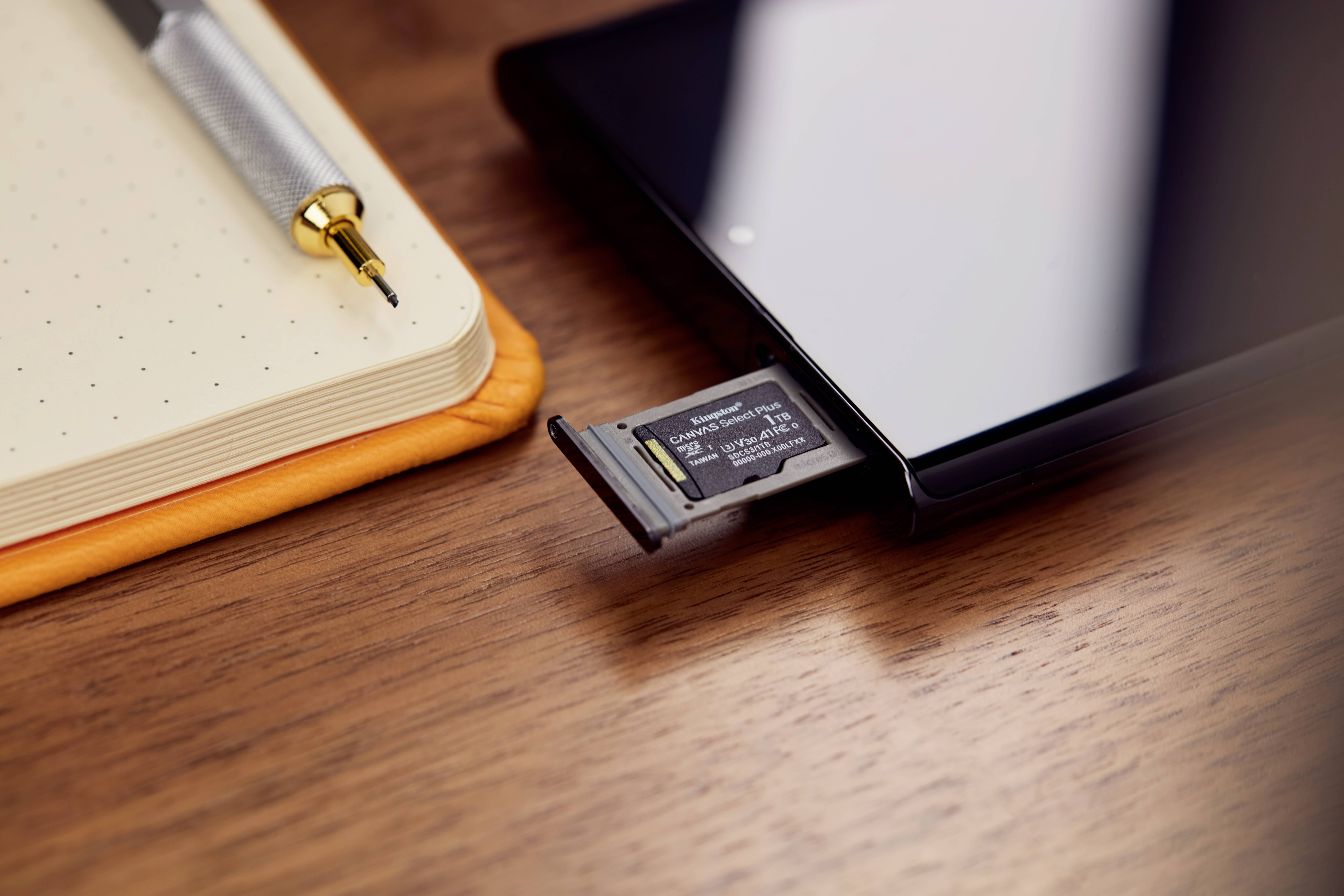 Eine microSD-Karte, die teilweise in ein Gerät auf einem Holztisch eingesteckt ist, neben einem Stylus und einem Notizbuch.