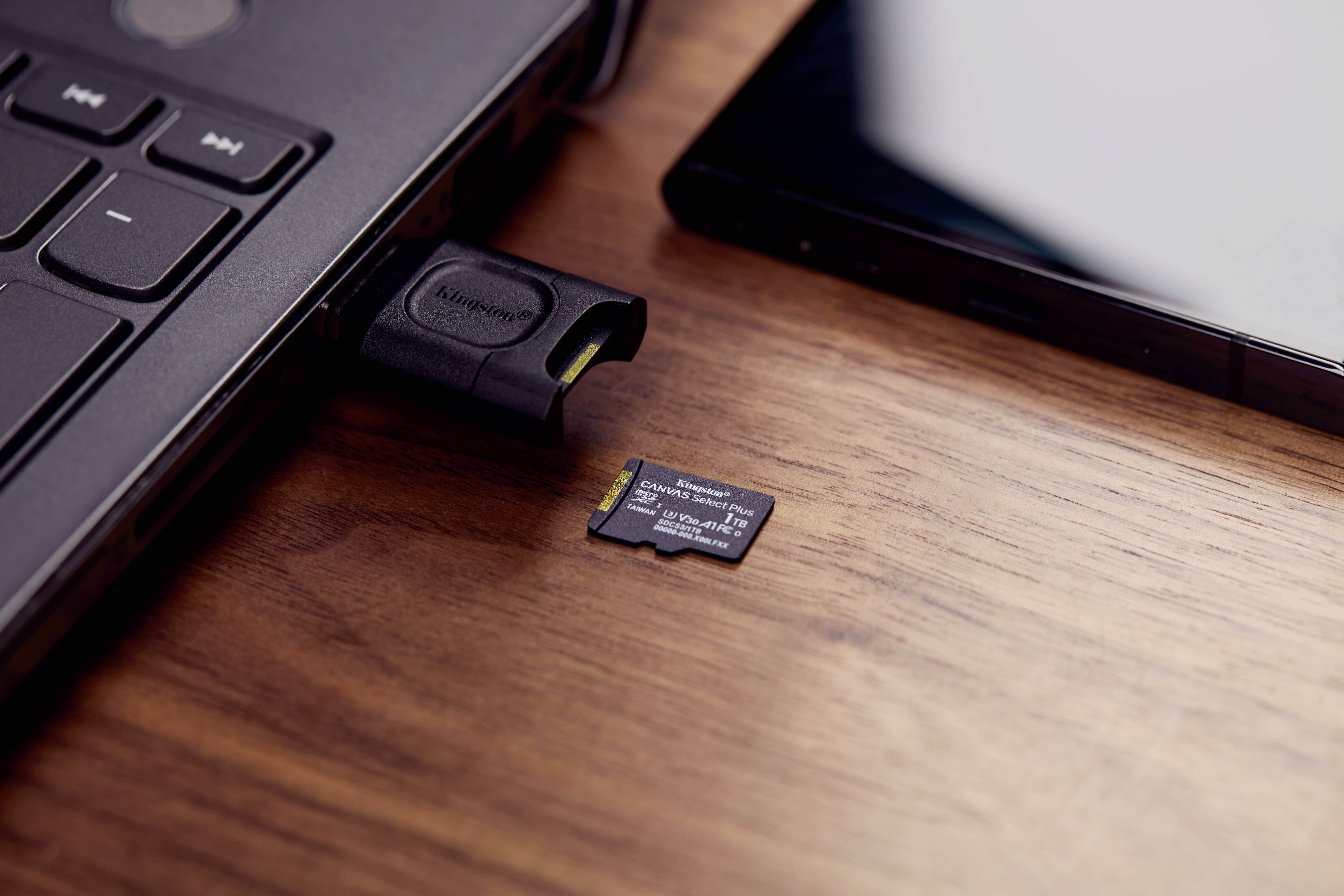 Ein Laptop mit einem USB-Kartenleser, der eingesteckt ist, mit einer microSD-Karte daneben auf einer Holzoberfläche und einem Smartphone in der Nähe.