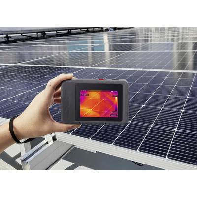 Eine Person hält ein Wärmebildgerät, das eine Wärmekarte von Solarpanels zeigt, während die Panels unter einem klaren Himmel zu sehen sind.