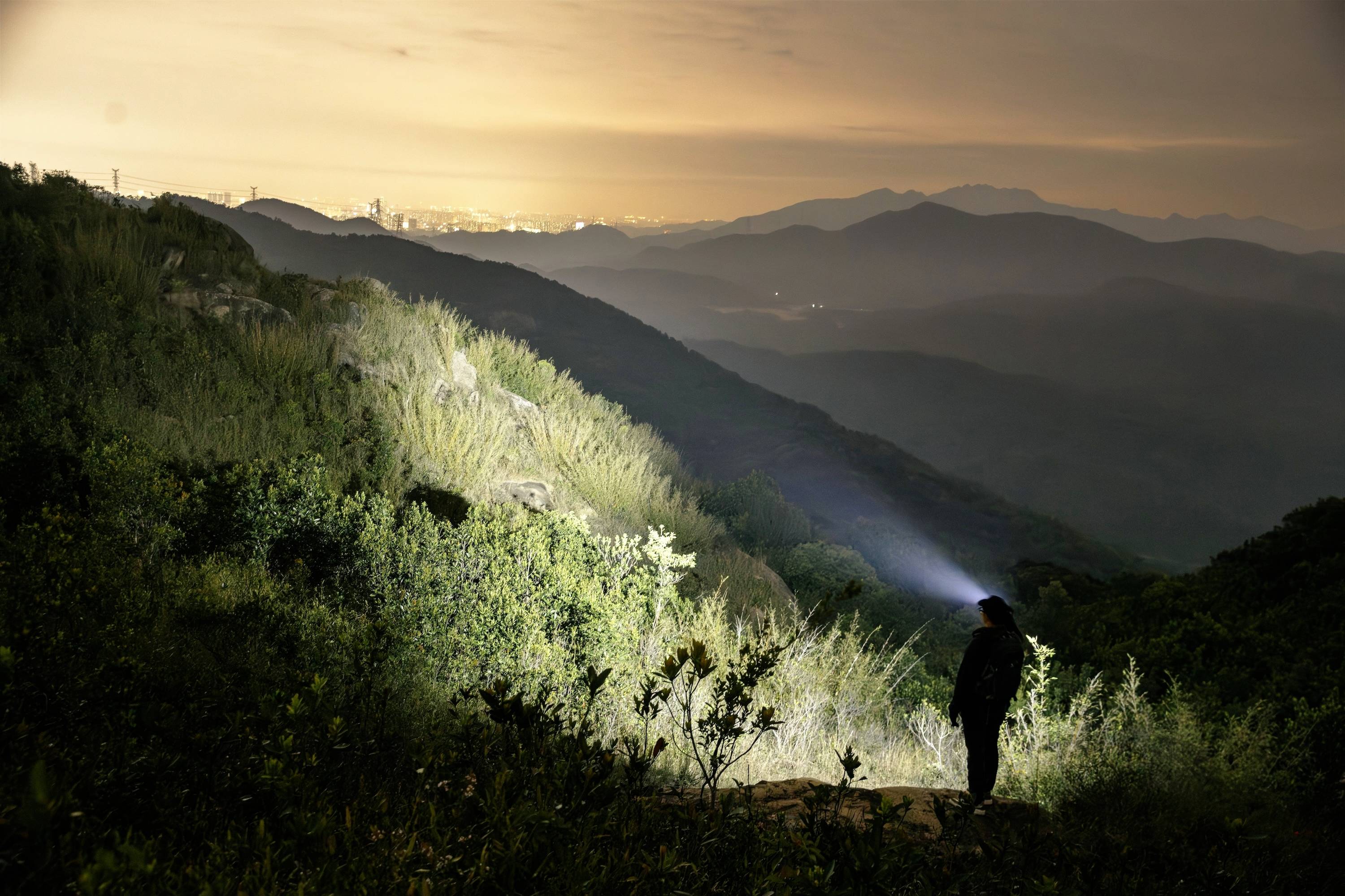 Eine Person mit einer Taschenlampe steht auf einem Hügel in der Nacht und blickt auf eine ferne Stadt, die von orangefarbenen Lichtern beleuchtet wird, mit Bergen im Hintergrund.