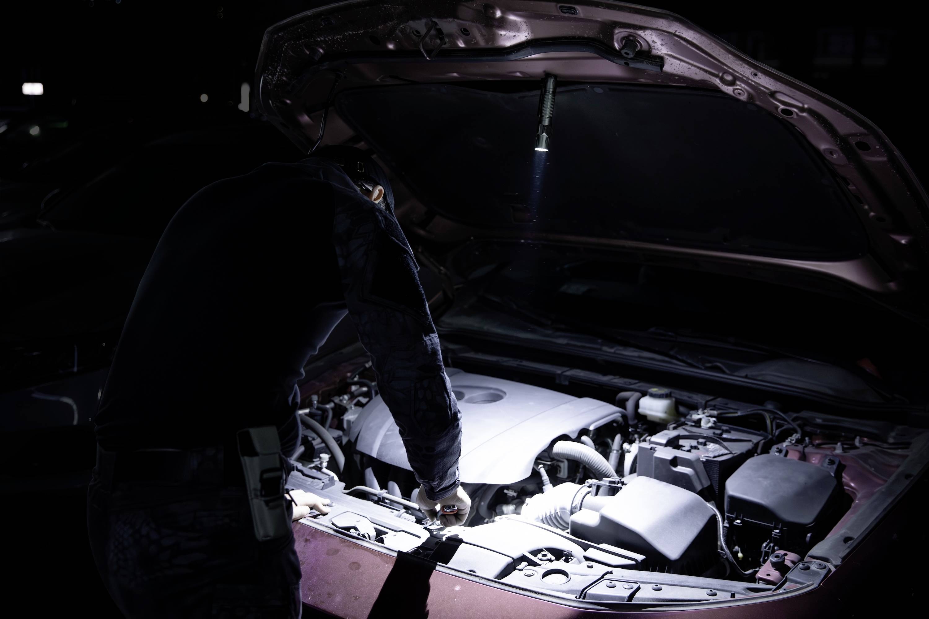 Eine Person in dunkler Kleidung untersucht bei Nacht unter einer offenen Motorhaube einen Automotor, wobei sie sich mit einer Taschenlampe beleuchtet.