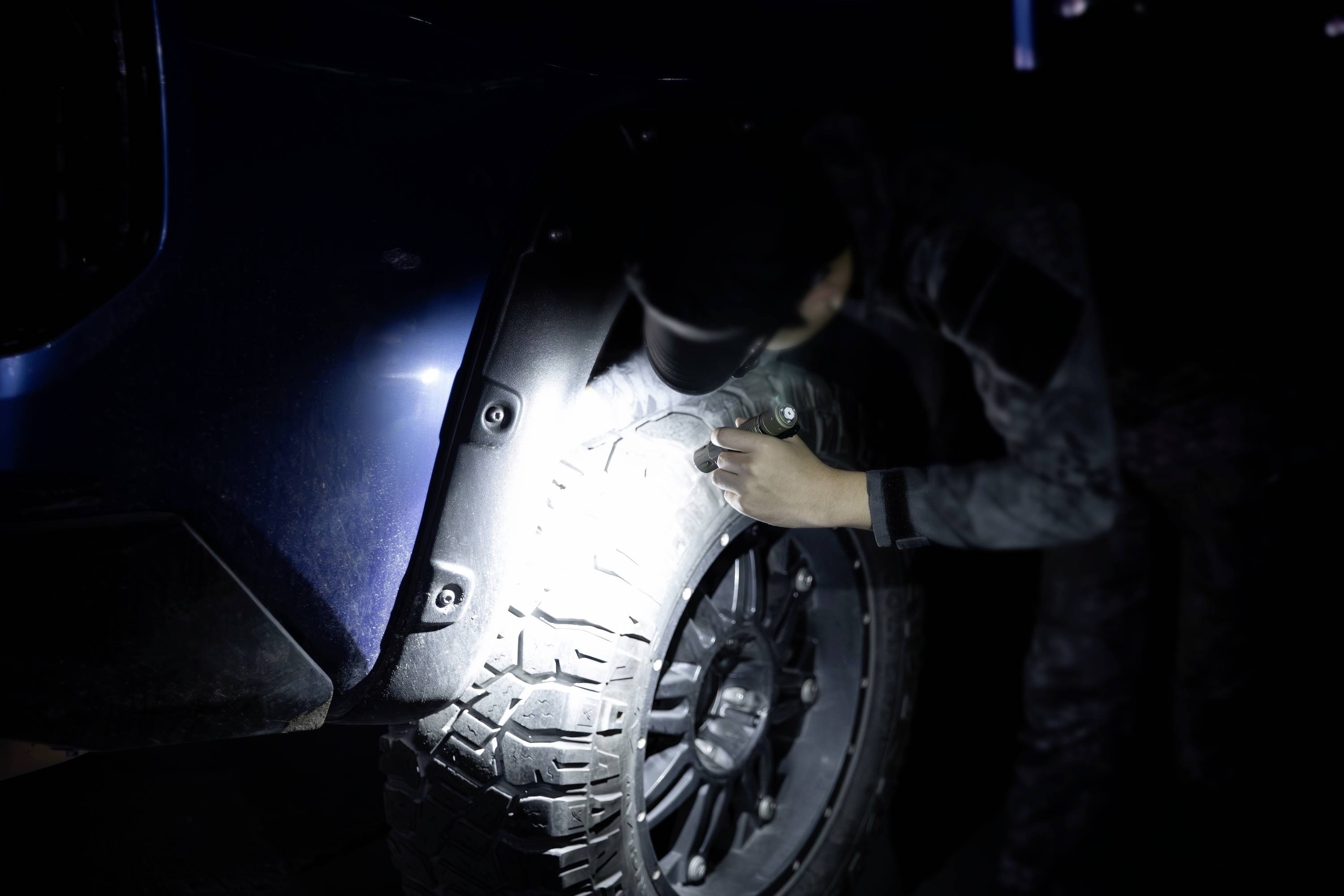 Eine Person, die bei Nacht mit einer Taschenlampe einen Autoreifen inspiziert und sich dabei auf das Profil des Reifens konzentriert, wobei die Notwendigkeit von Sicherheits- und Wartungschecks hervorgehoben wird.