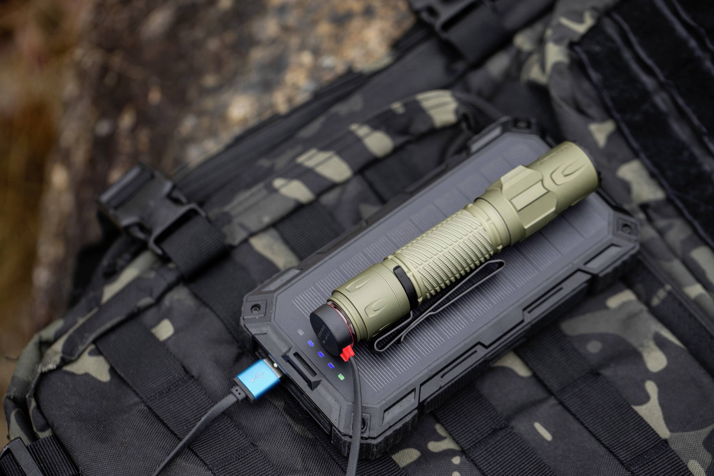 Drei Taschenlampen in Grün, Schwarz und Blau sind an der Seite eines getarnten Rucksacks im Freien befestigt und zeigen ihre Größen und Farben.