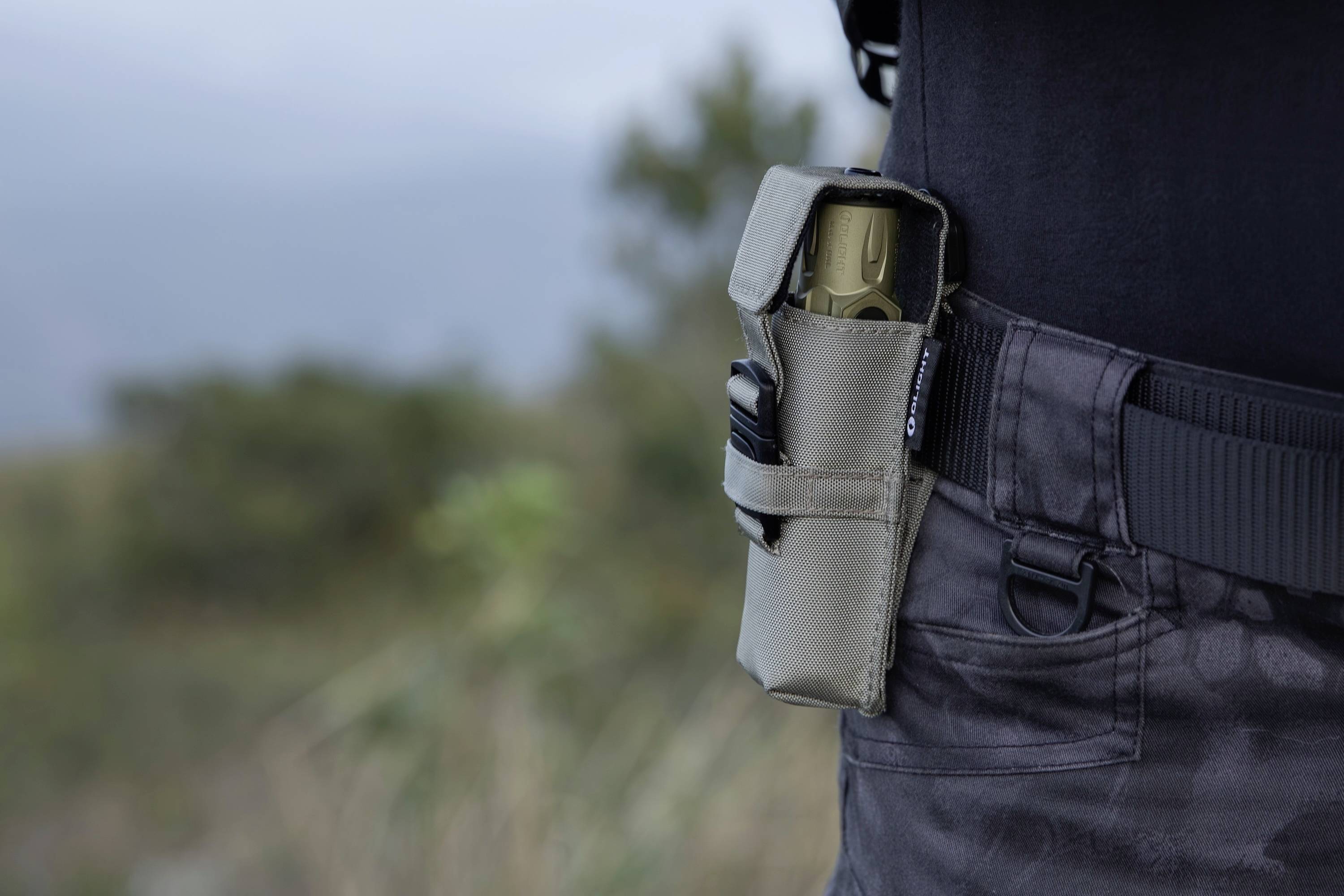 Eine Person trägt dunkle taktische Hosen und ein schwarzes Hemd und hat eine Tasche an der Hüfte, in der ein Multitool oder ein ähnliches Utensil verstaut ist, im Freien.
