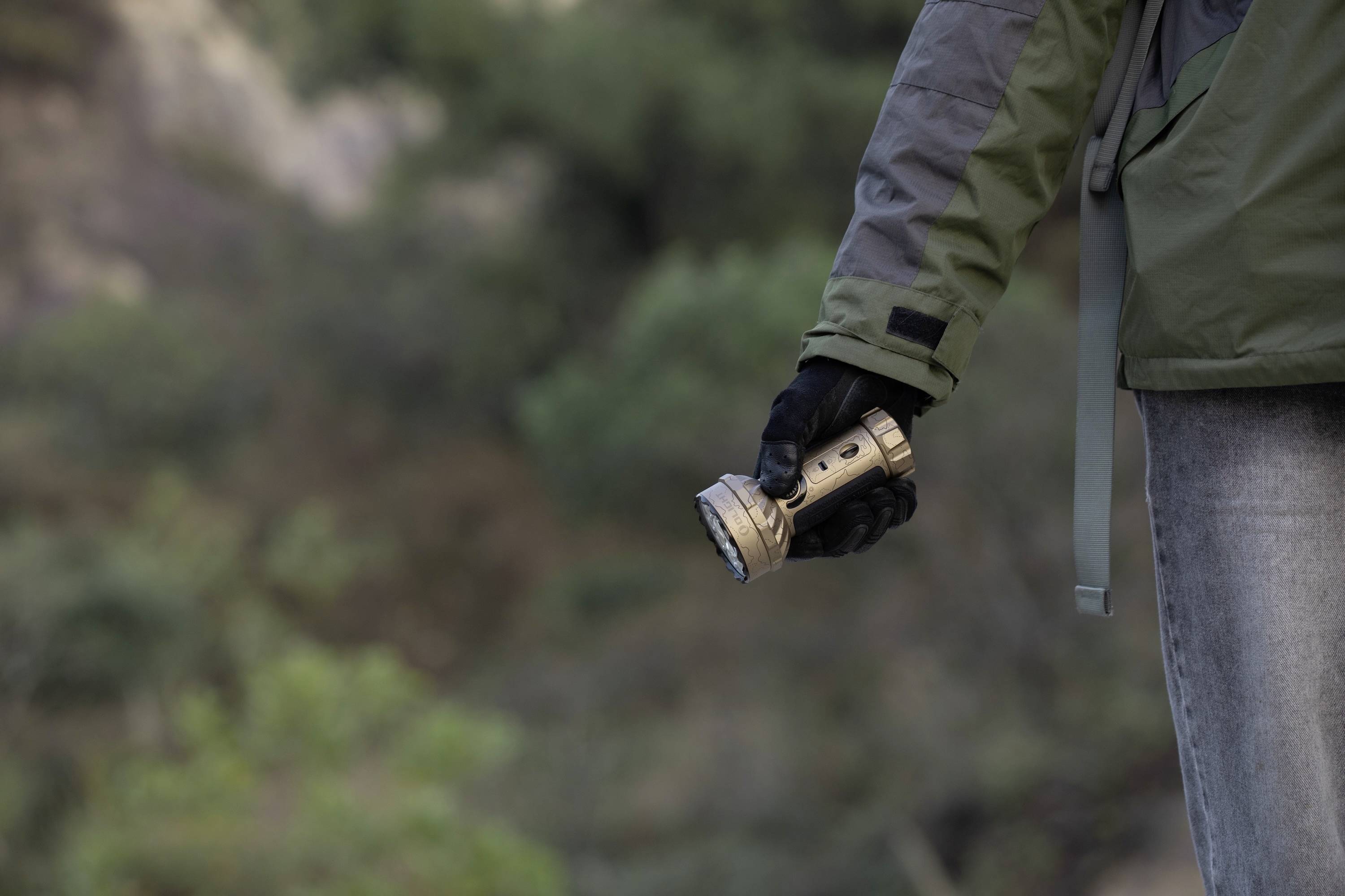 Eine Person, die eine Taschenlampe in einer Hand hält, trägt eine grüne Jacke und schwarze Handschuhe. Sie steht im Freien mit verschwommener Vegetation im Hintergrund.