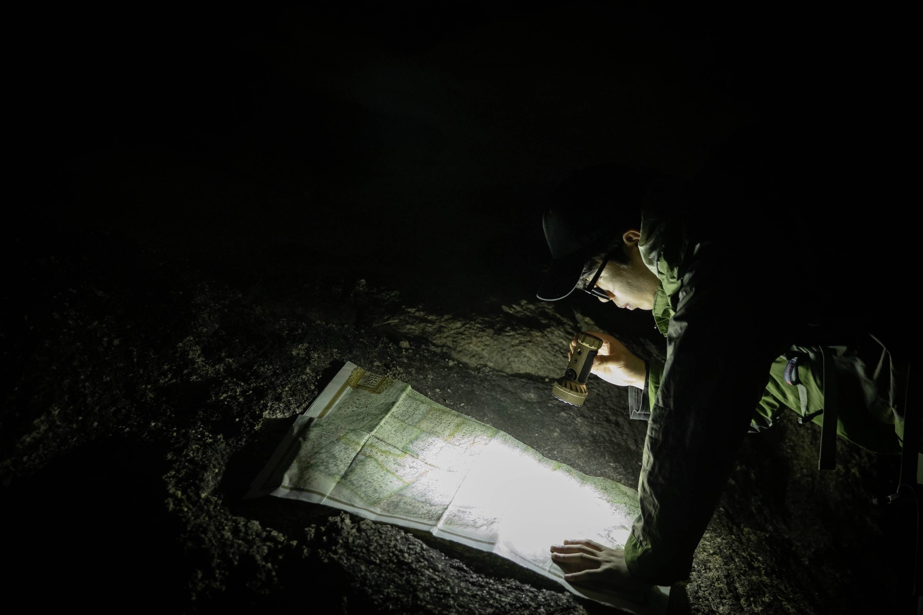 Eine Person beugt sich über eine Karte in der Dunkelheit, von einer Taschenlampe beleuchtet, was Navigation oder Erkundung in einer dunklen Umgebung andeutet.