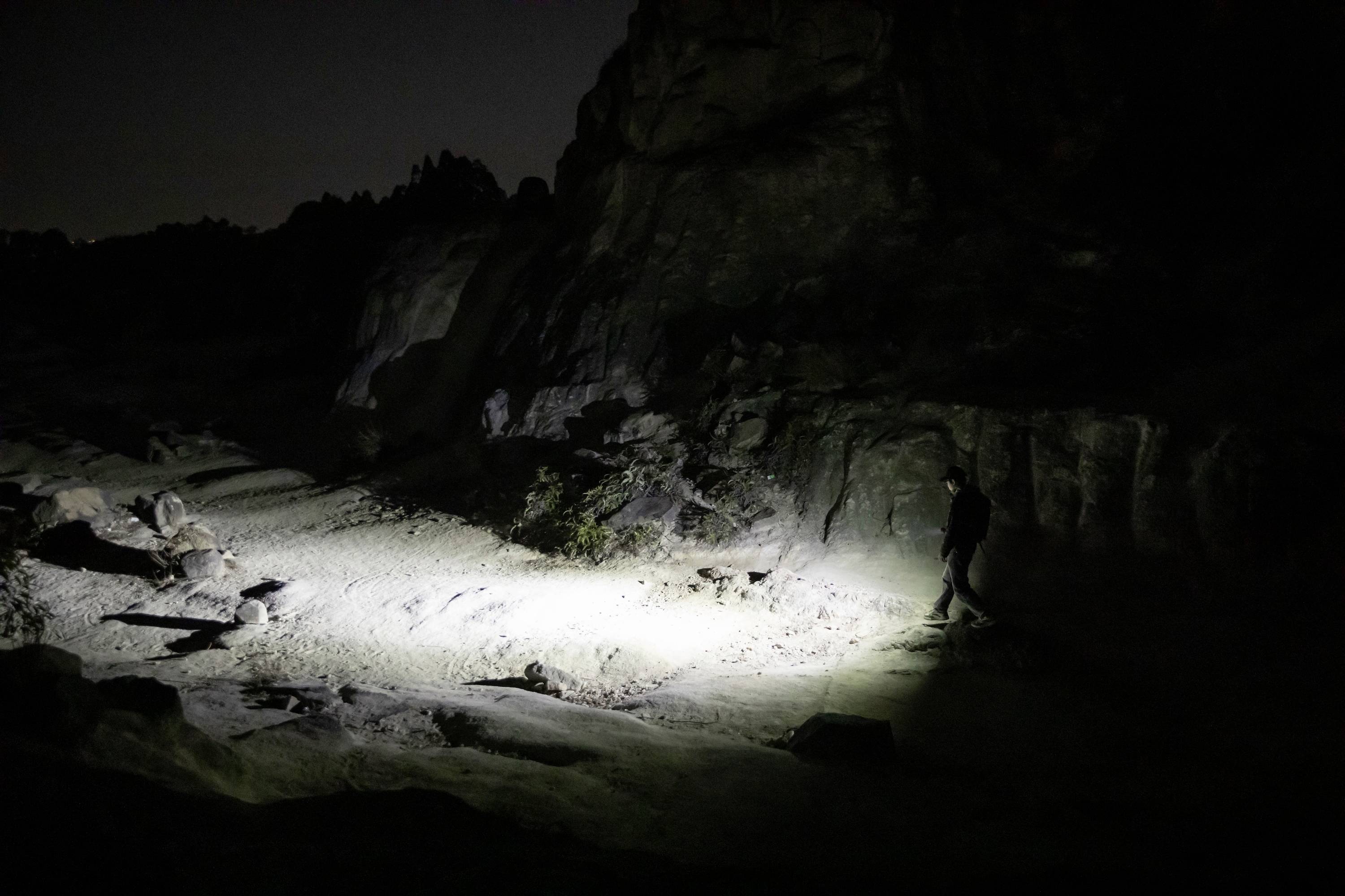 Eine Person geht nachts auf einem felsigen Pfad, wobei eine Taschenlampe den Boden beleuchtet, umgeben von dunkler, rauer Landschaft.