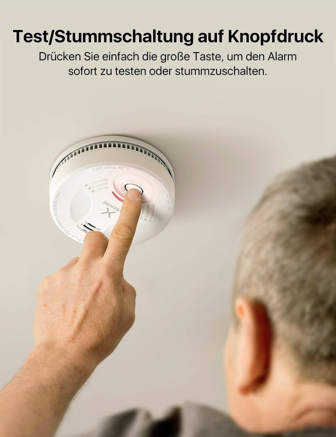 Ein Mann drückt eine Taste auf einem Rauchmelder, um den Alarm zu testen oder stummzuschalten. Text: 'Test/Stummschaltung auf Knopfdruck'.